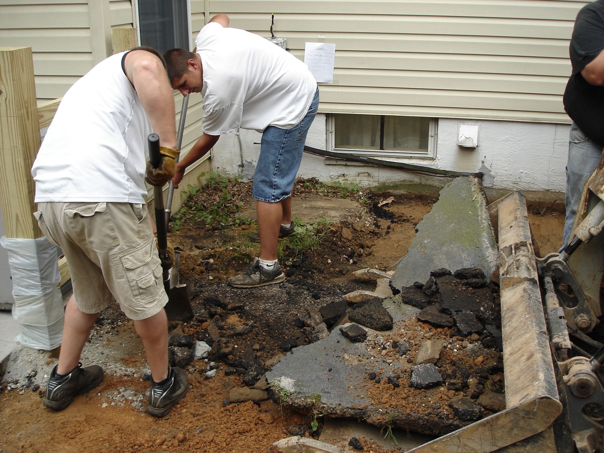 Two people in white shirts and shorts dig into the dirt in front of a house, clearing away chunks of broken concrete.