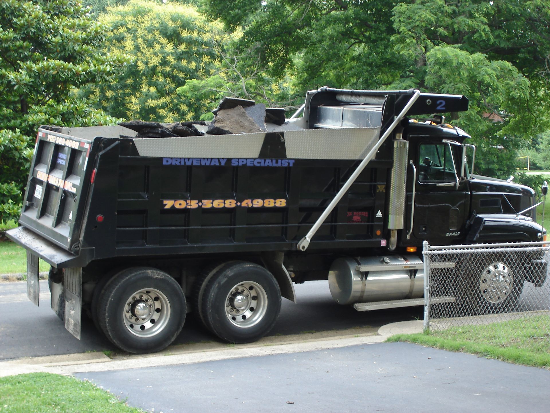 A black dump truck loaded with debris parked on a paved driveway near a chain-link fence and green trees.