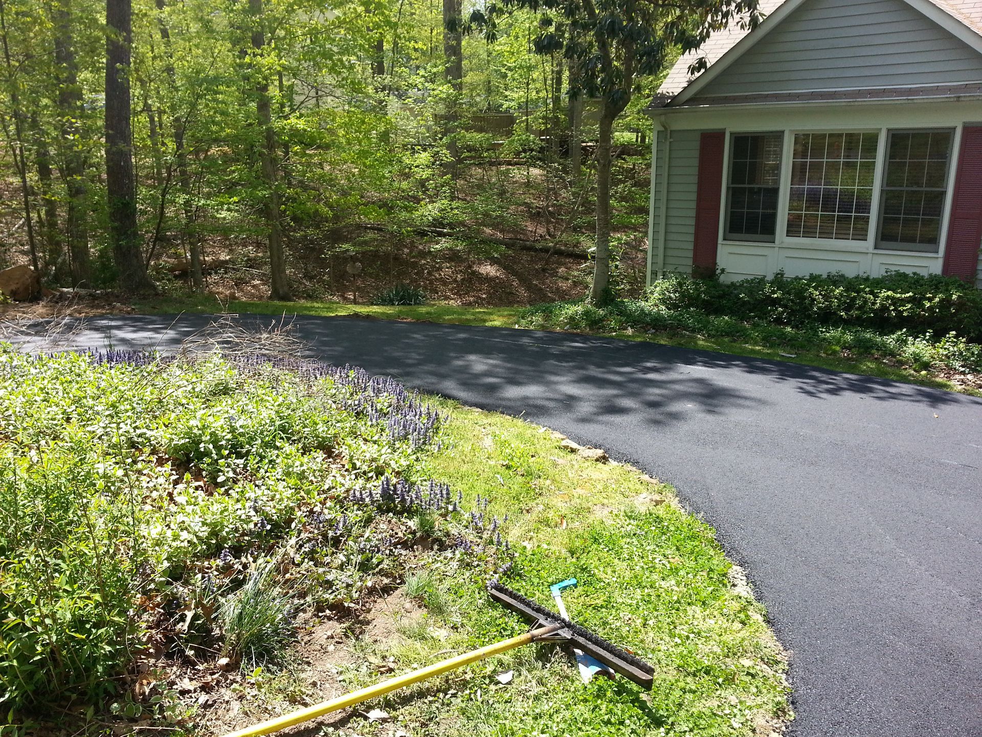 A newly paved asphalt driveway curves past a house with a yellow tool resting on the grass in the foreground.