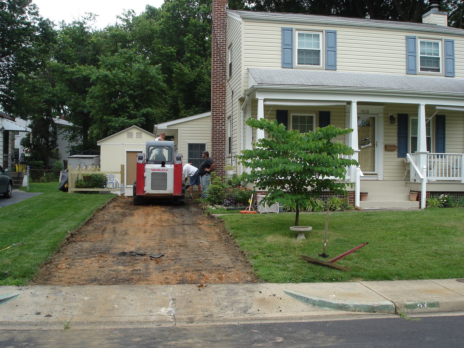 A construction vehicle clears dirt for a new driveway in front of a two-story house with blue shutters and a porch.