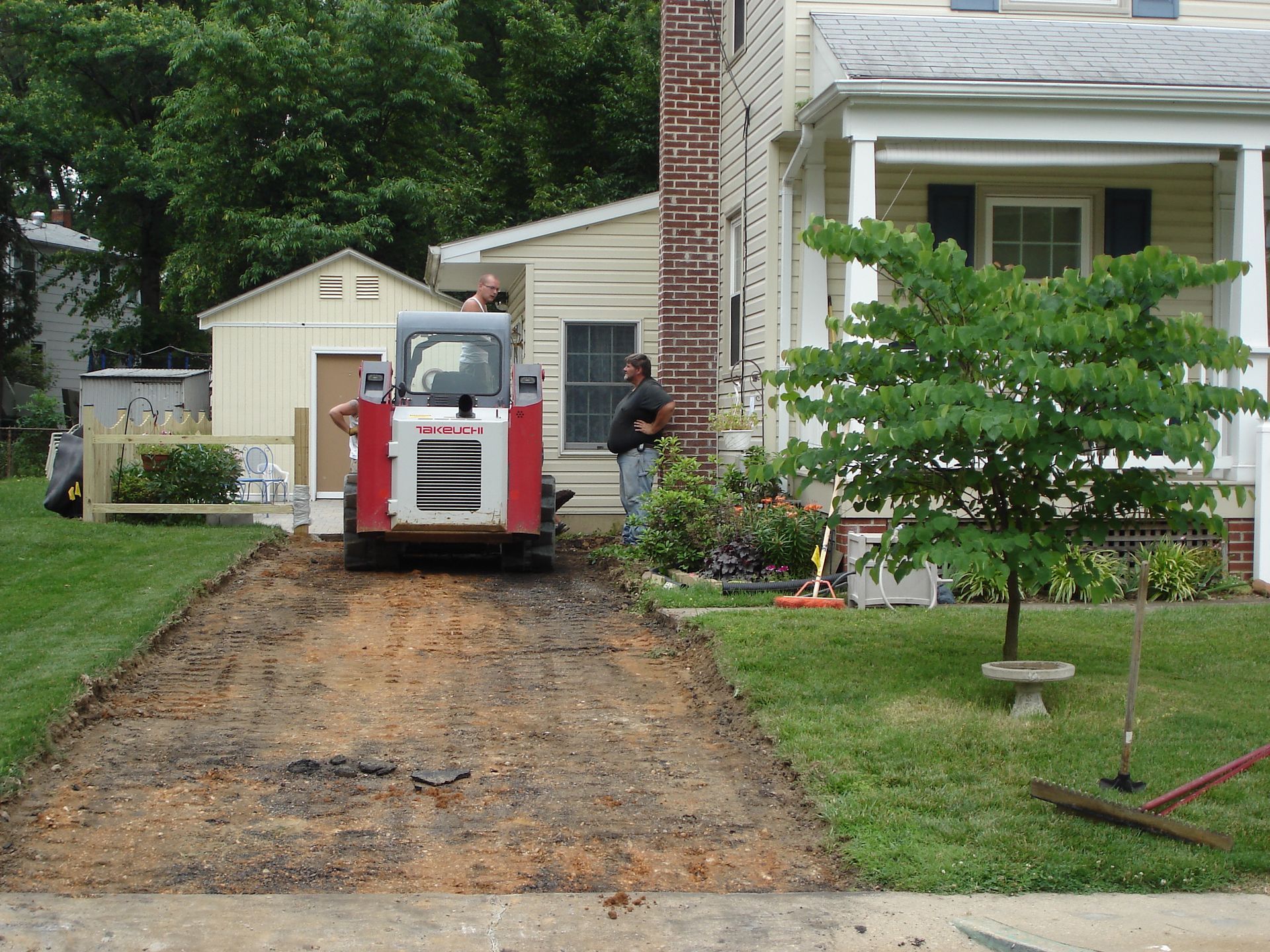 A skid steer sits in a dug-out driveway next to a house, with two workers standing nearby in a suburban yard.