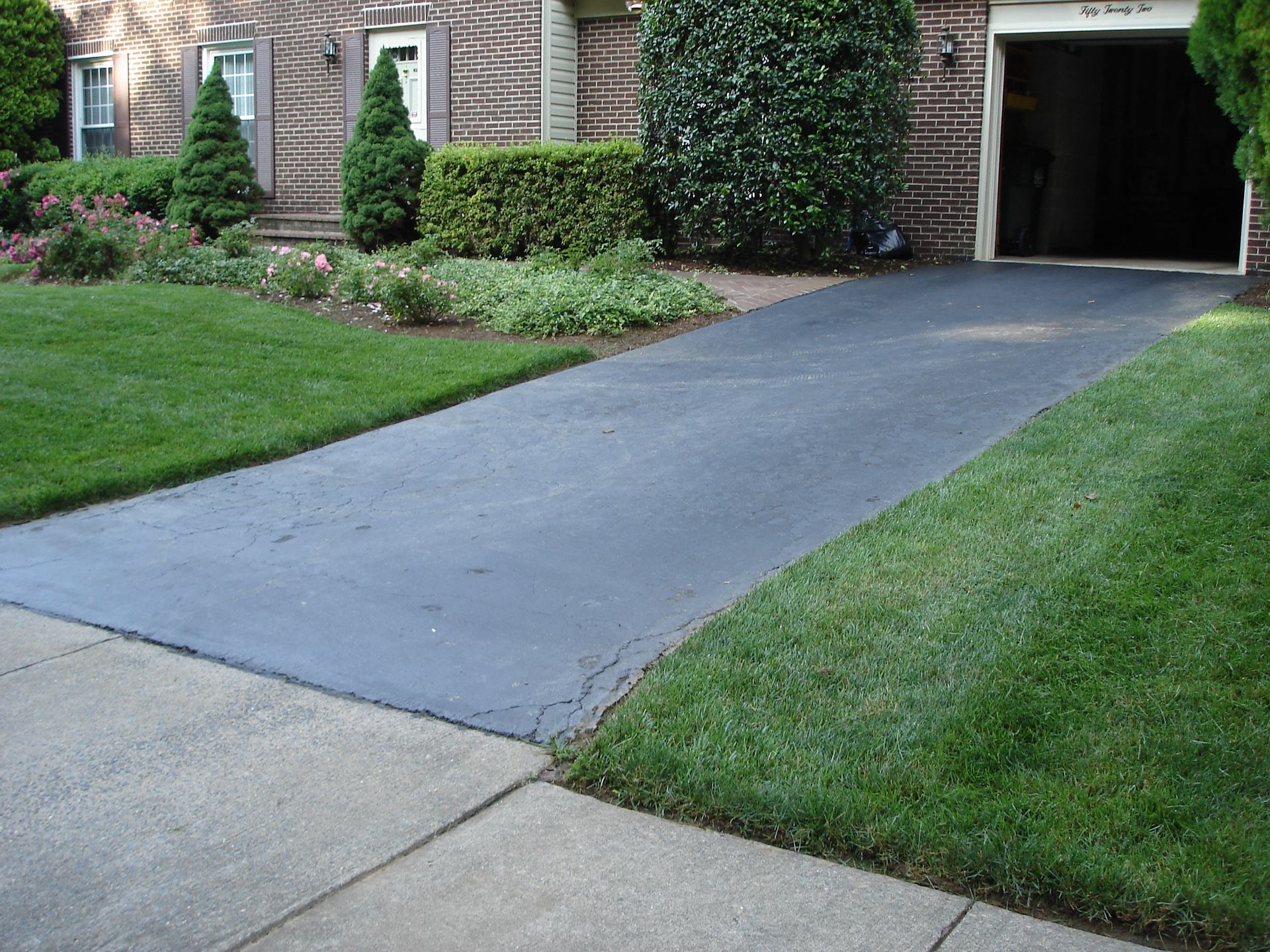 A brick house with a dark asphalt driveway extending from the street to an open garage, flanked by lush green lawns.