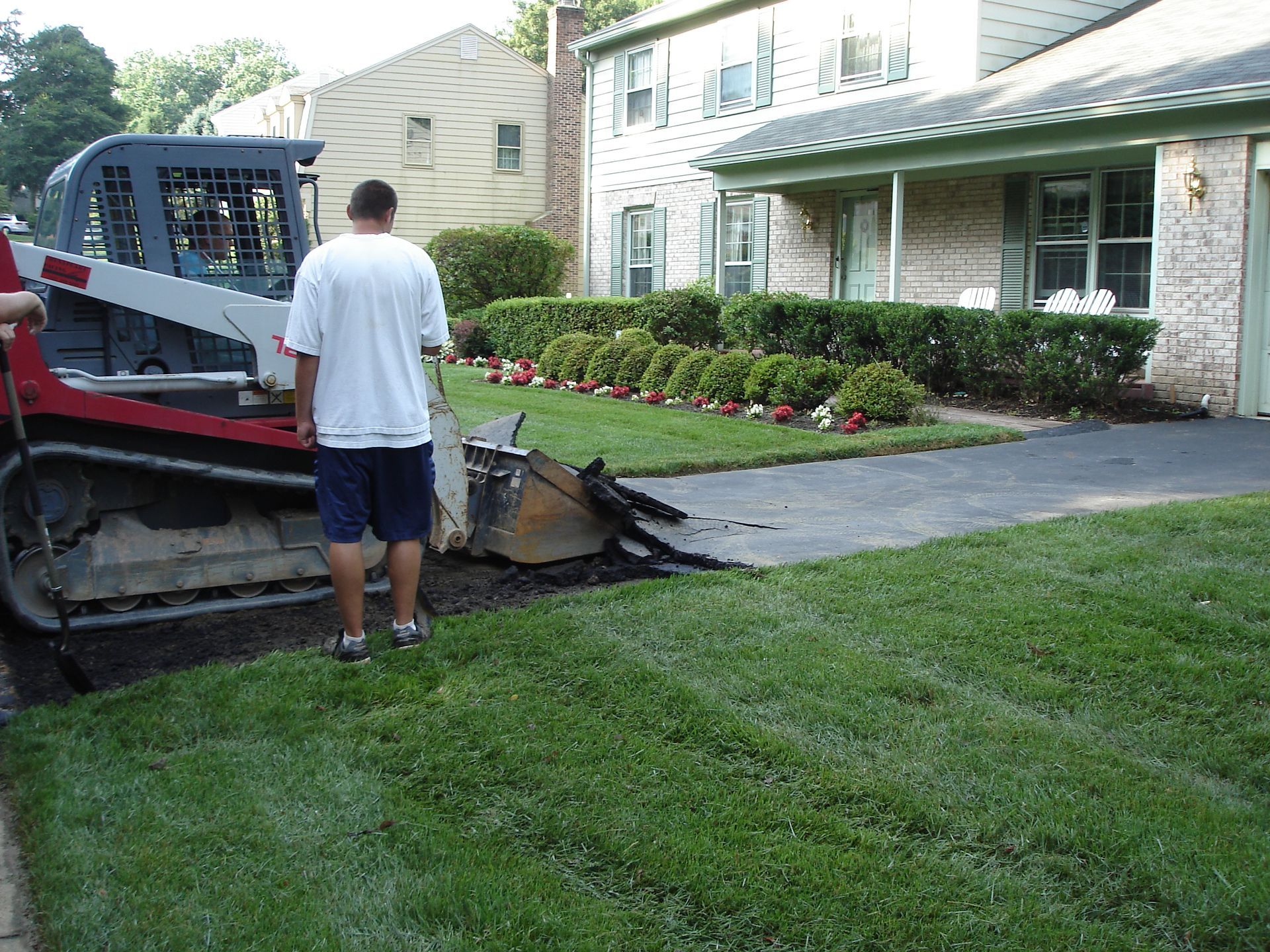 A person operates a skid-steer loader to remove pavement from a residential driveway in front of a house.