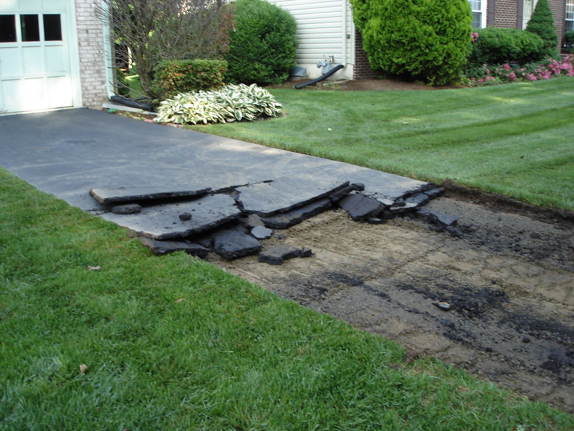 A residential asphalt driveway with a section of broken, crumbling pavement and exposed dirt near the lawn.