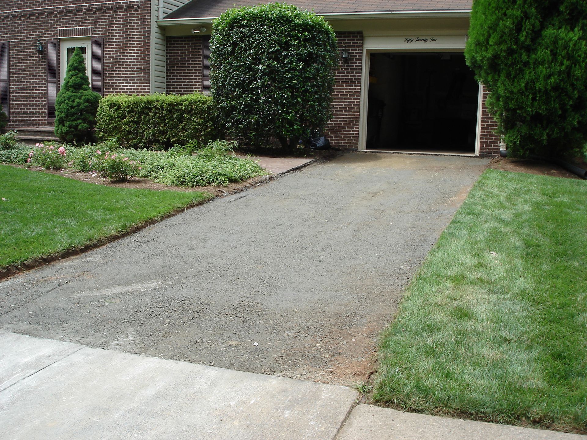 A gravel driveway leading from a paved street toward a residential house garage, framed by lush green lawn and bushes.