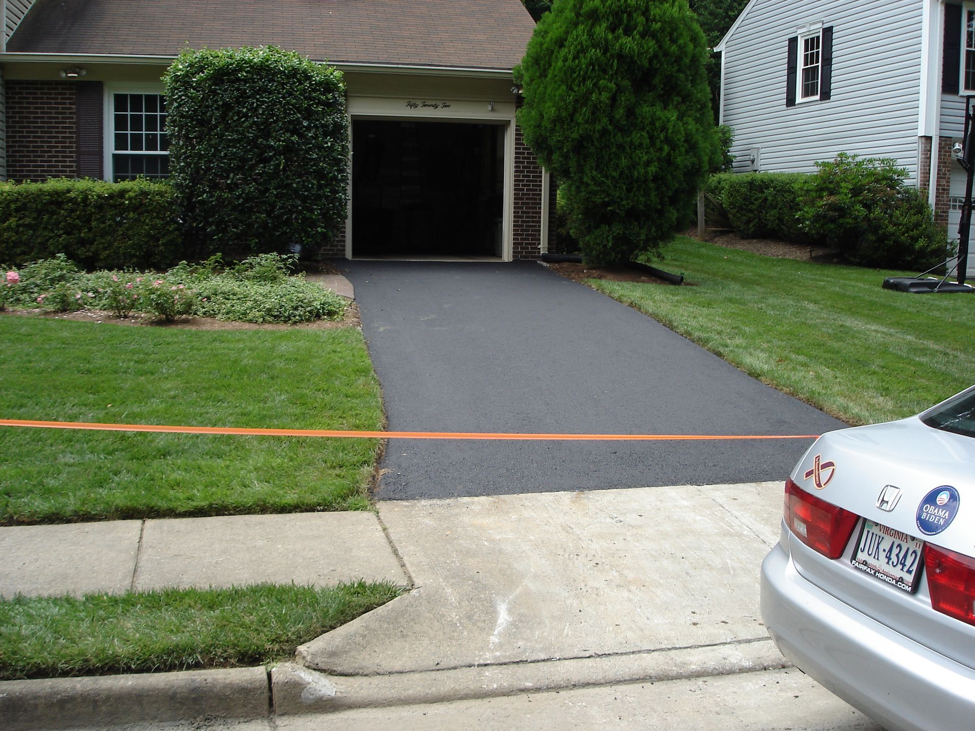 A silver car is parked on the street next to a residential driveway that has been freshly paved with new black asphalt.