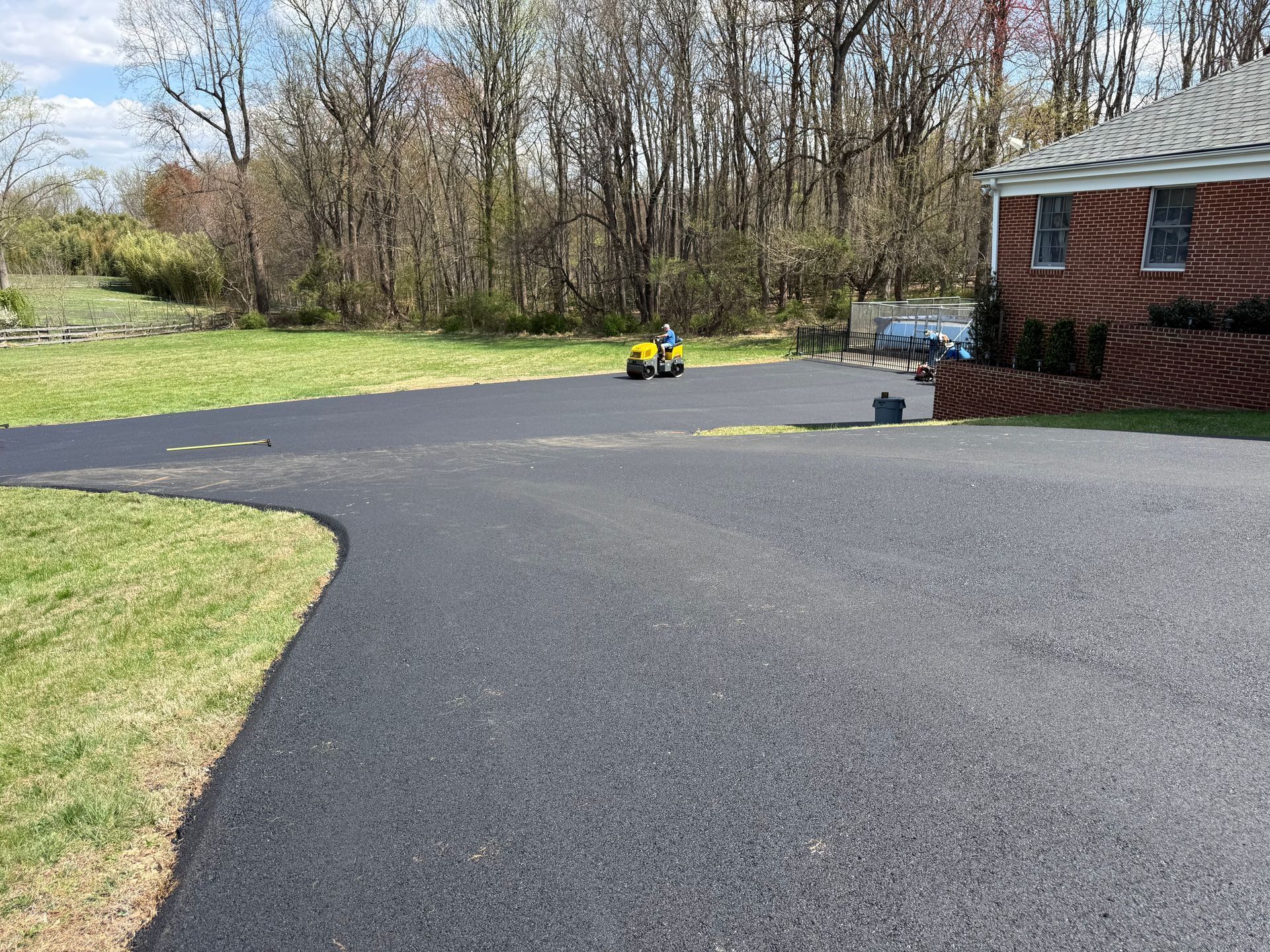 A person on a yellow riding mower cuts grass beside a newly paved, smooth black asphalt driveway next to a brick house. 