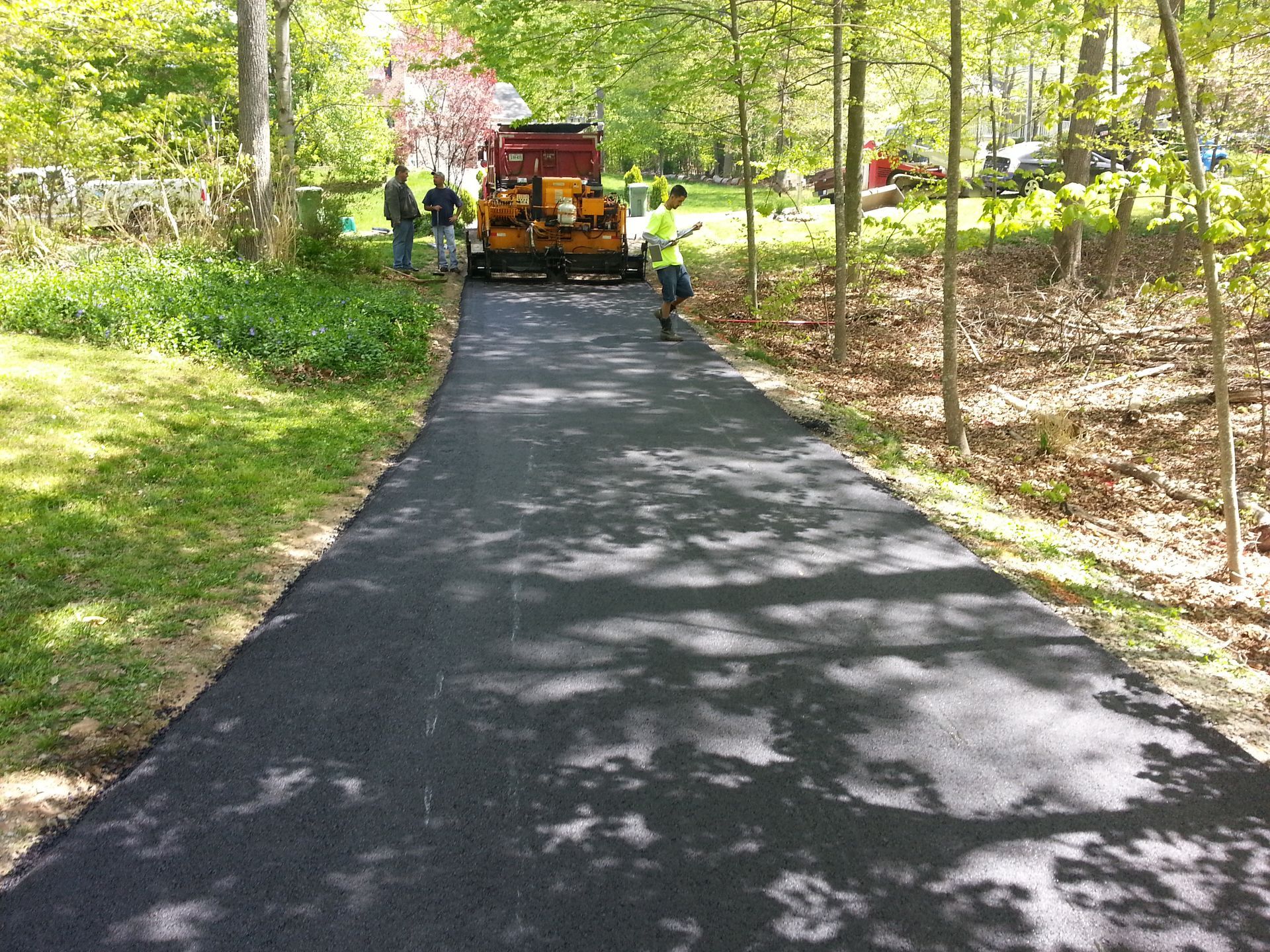 A construction crew paves a driveway with black asphalt in a wooded residential area.