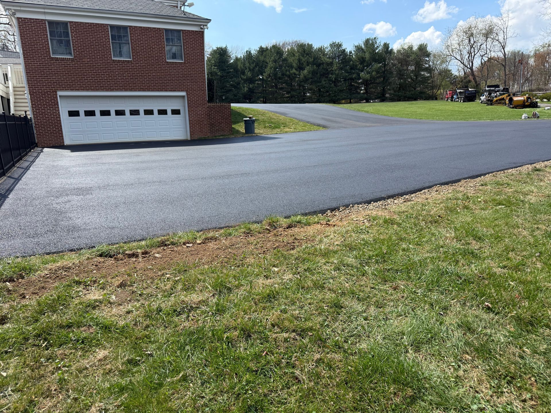 A newly paved dark asphalt driveway leads to a brick house with a white garage door, set against a grassy lawn and trees. 