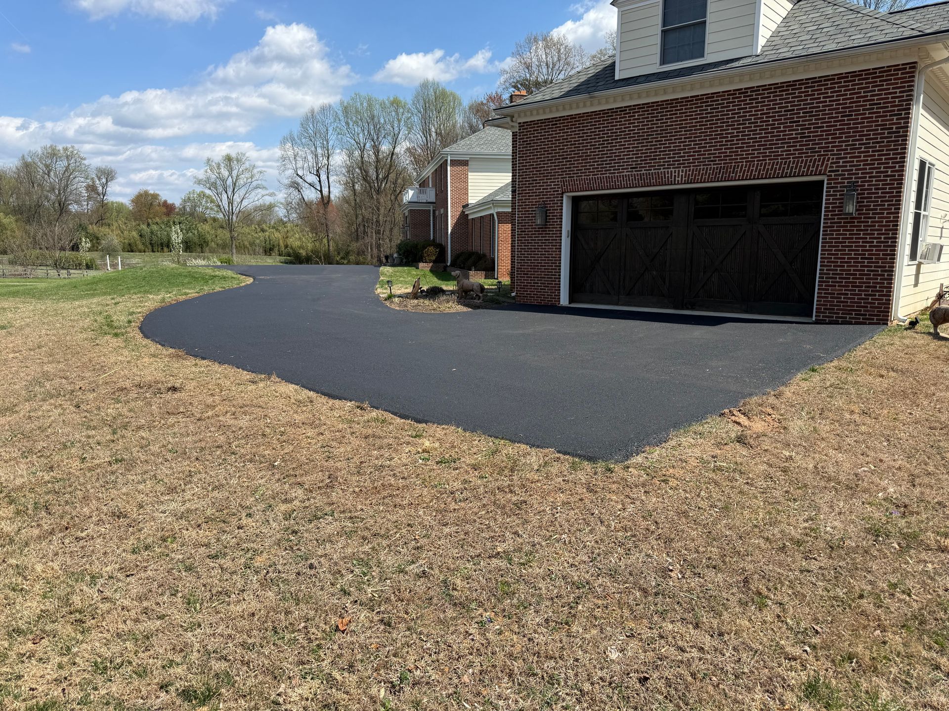 A freshly paved black asphalt driveway leads to a brick garage attached to a house under a clear, blue sky. 