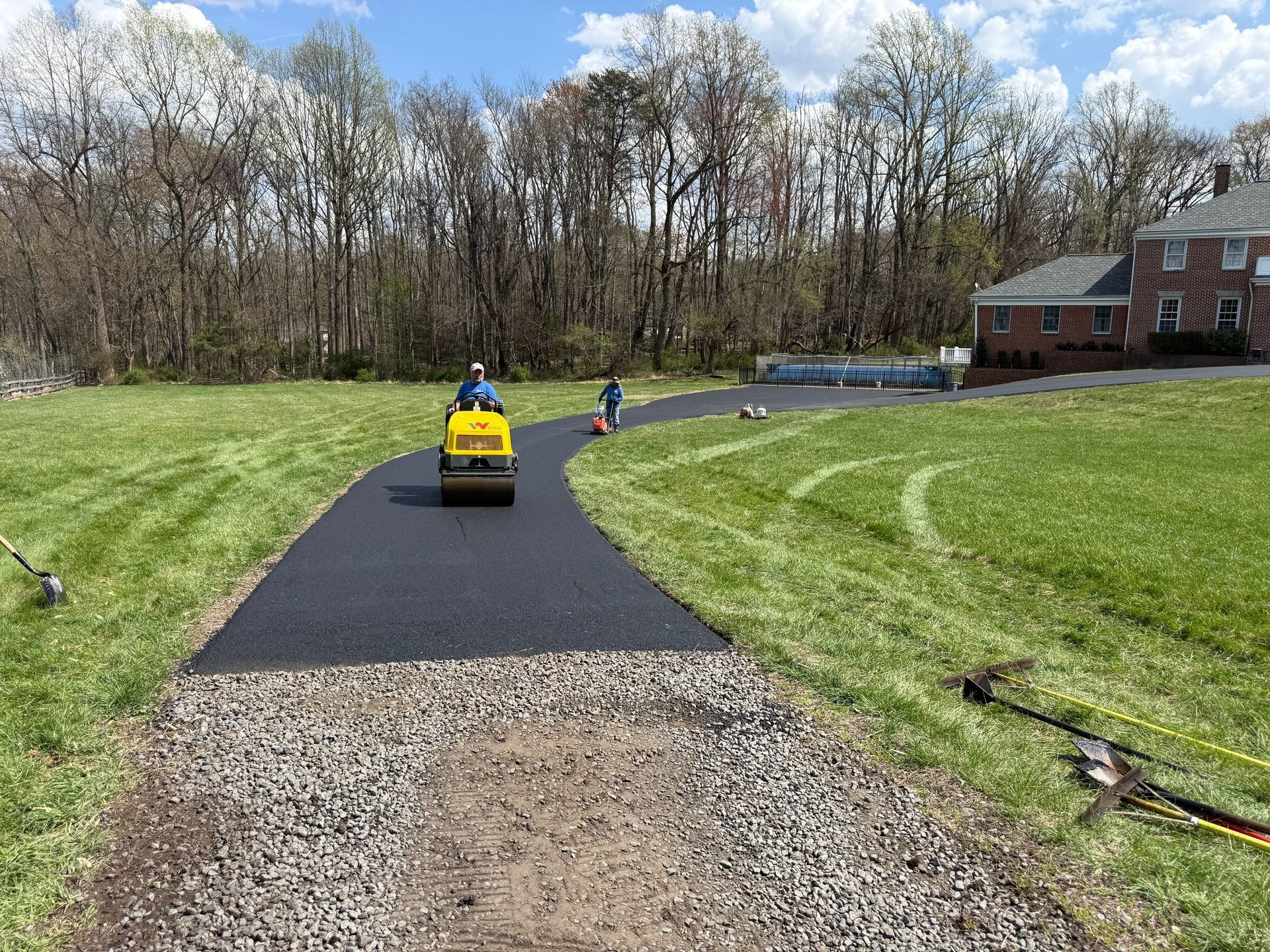A person drives a yellow asphalt roller over a freshly paved, dark driveway toward a brick house on a sunny day. 