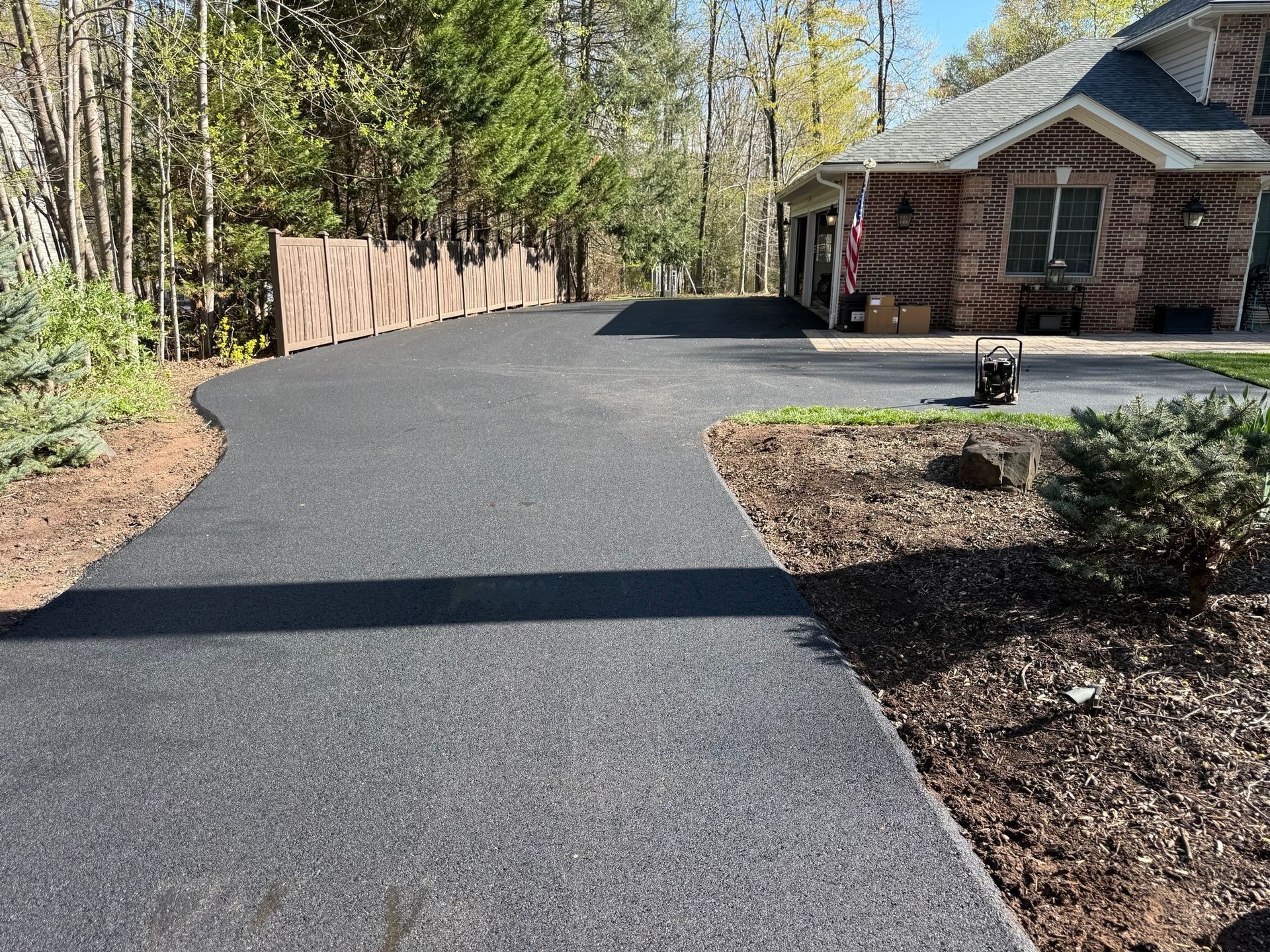 A freshly paved dark asphalt driveway leads toward a brick house surrounded by trees and a wooden fence.