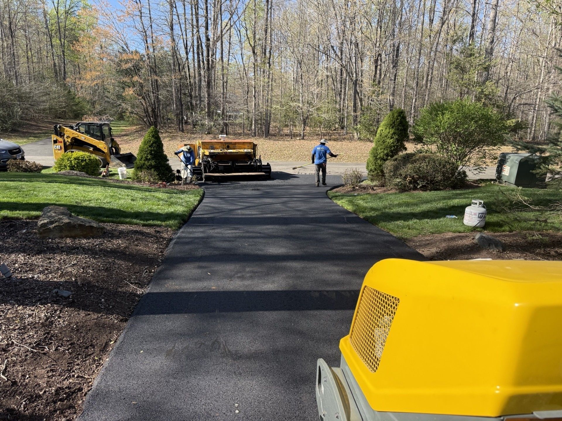 Construction workers operate machinery to pave a driveway with black asphalt in a residential outdoor setting.