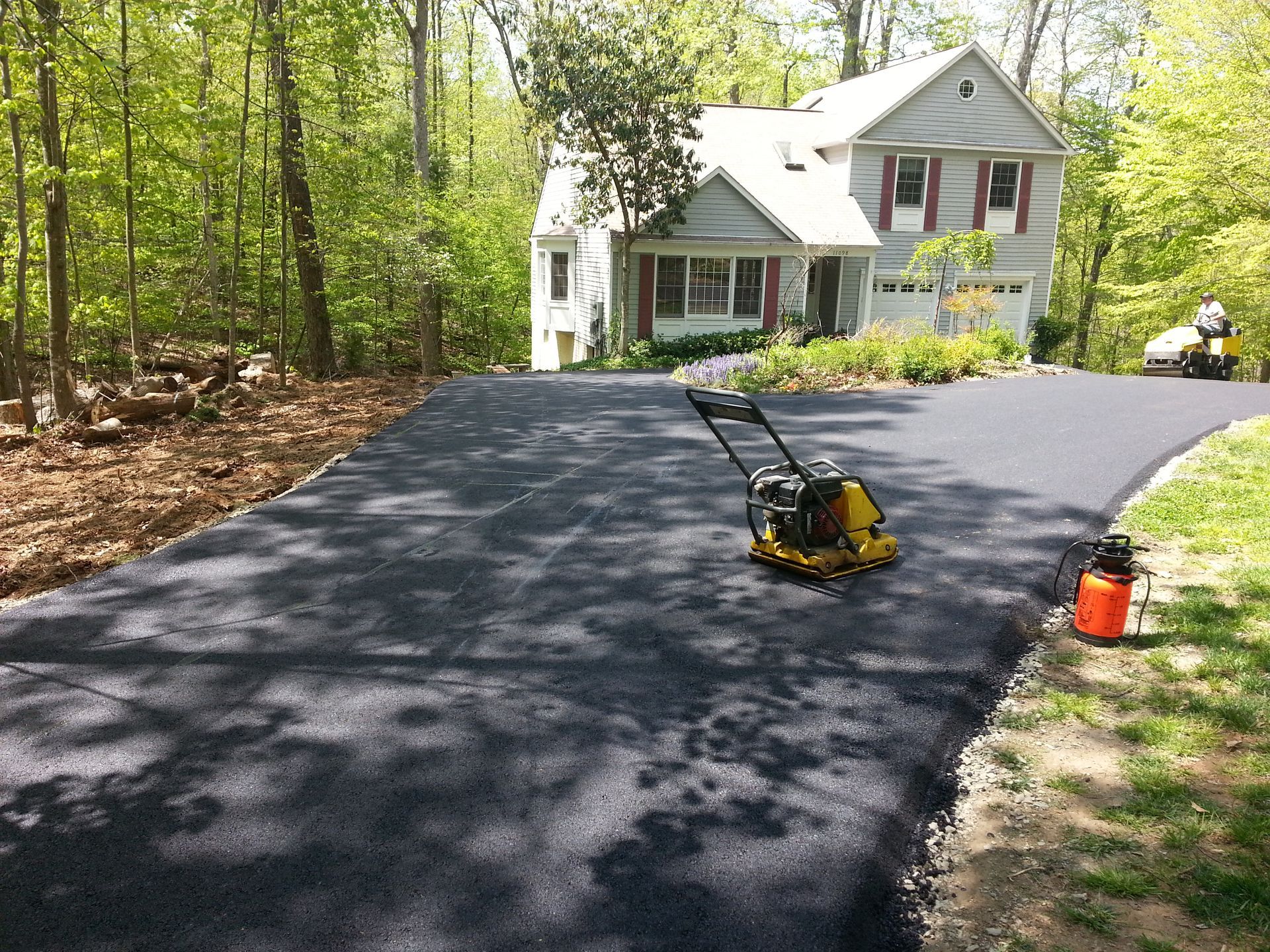 A yellow plate compactor sits on a newly paved black asphalt driveway leading to a house surrounded by trees.