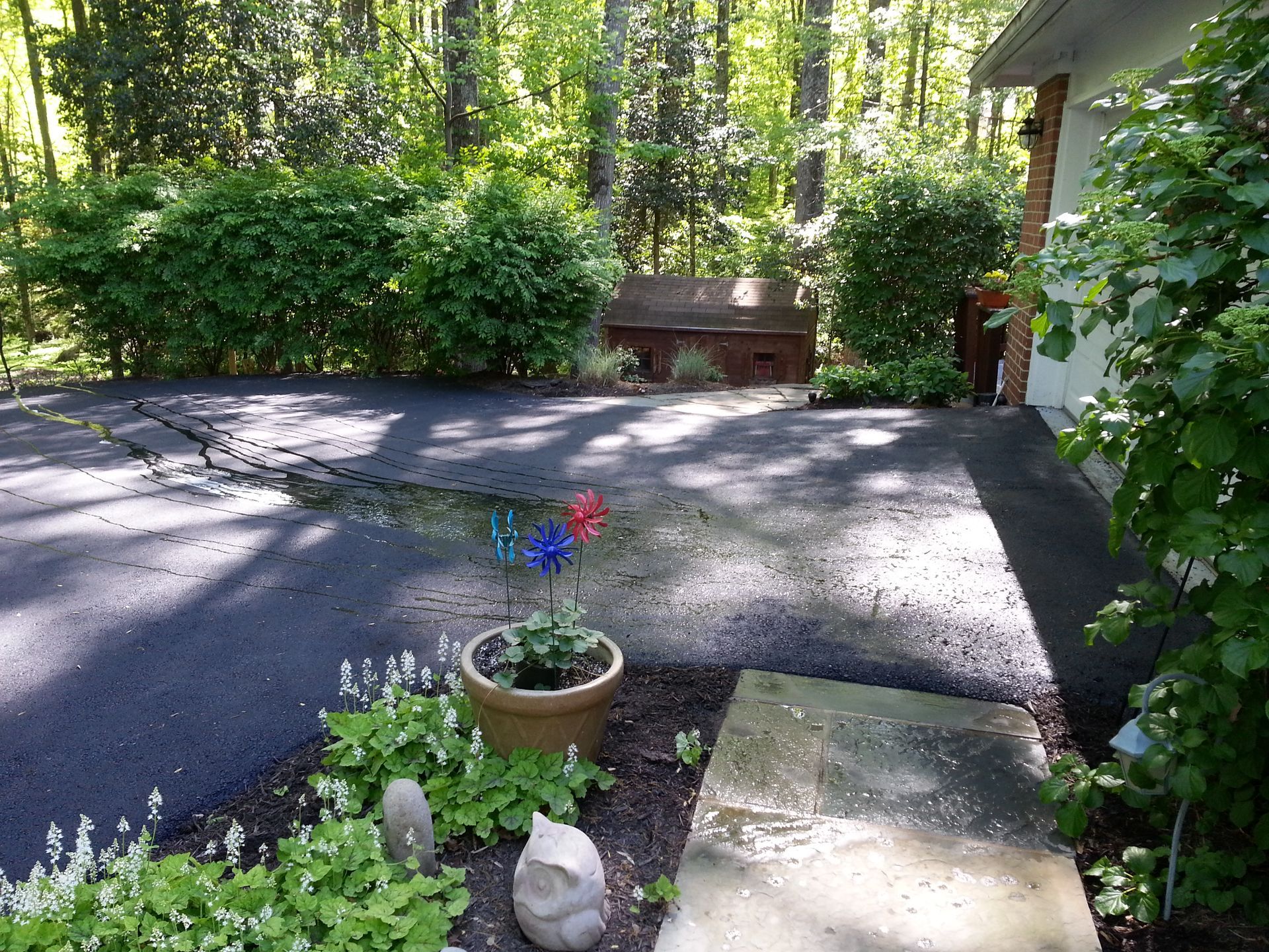 A dark, freshly paved driveway with a visible crack leads toward a wooded yard, with a potted flower and walkway in front.
