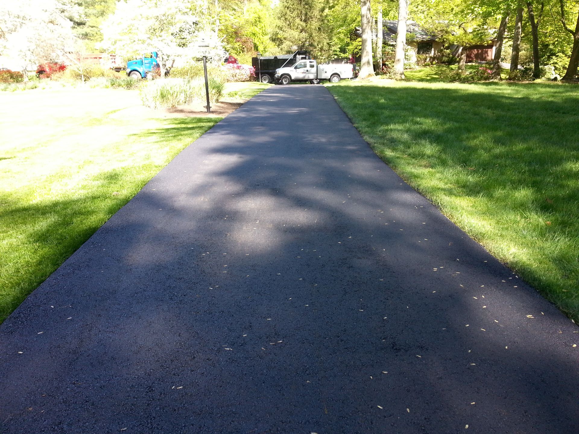 A freshly paved, dark asphalt driveway leading toward a house and parked vehicles, flanked by green grass on a sunny day.