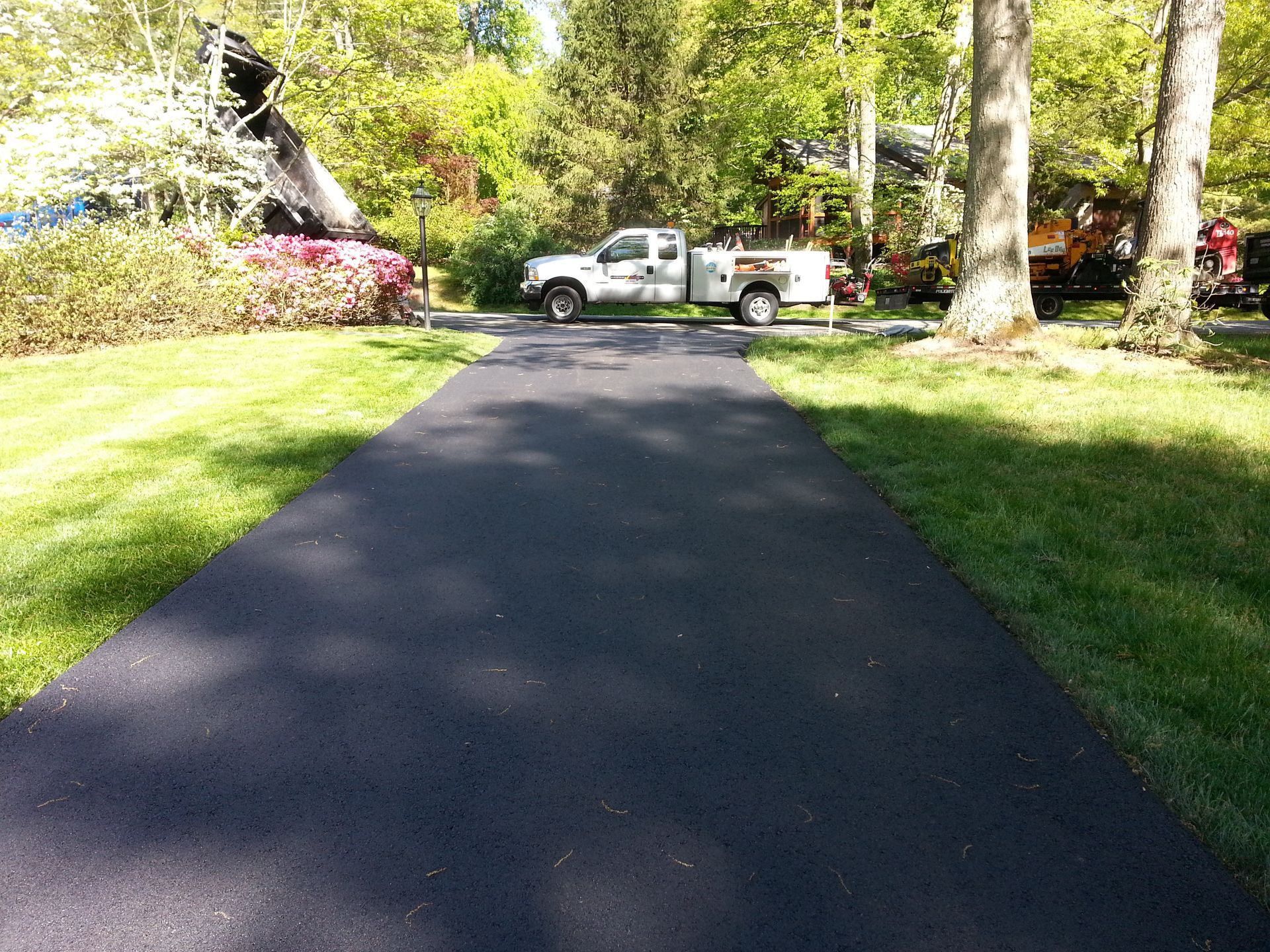 A freshly paved black asphalt driveway leads toward a white service truck parked in a sunny, green residential yard.