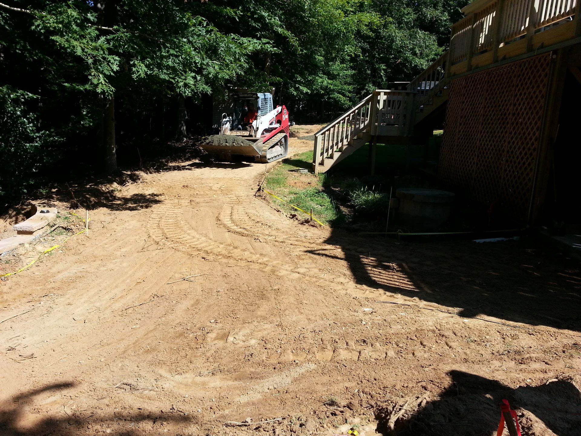 A red skid steer loader sits on a newly graded dirt path leading toward a house with a wooden deck.