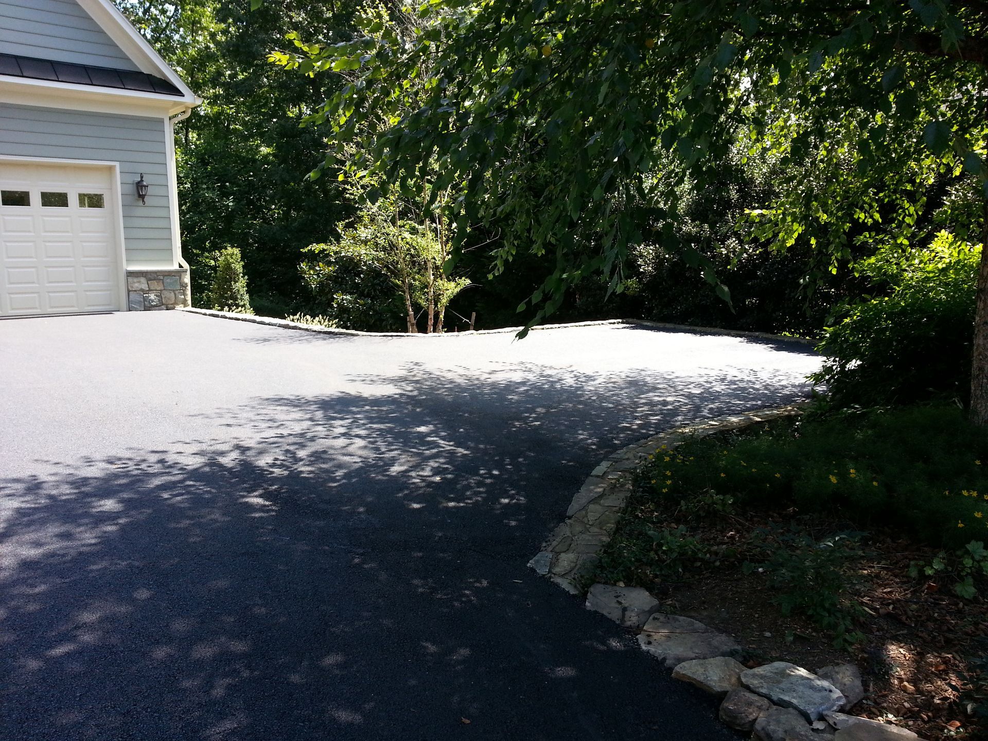 A light blue house with a white garage door, featuring a newly paved dark asphalt driveway bordered by natural stones.