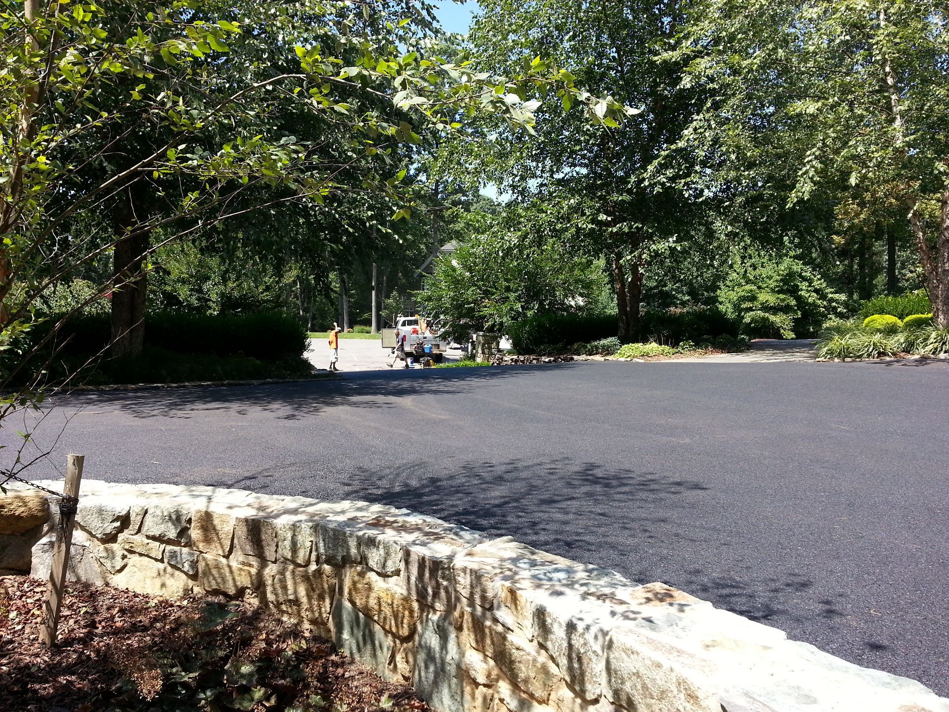 A freshly paved asphalt driveway leads toward a wooded area behind a decorative stone retaining wall.