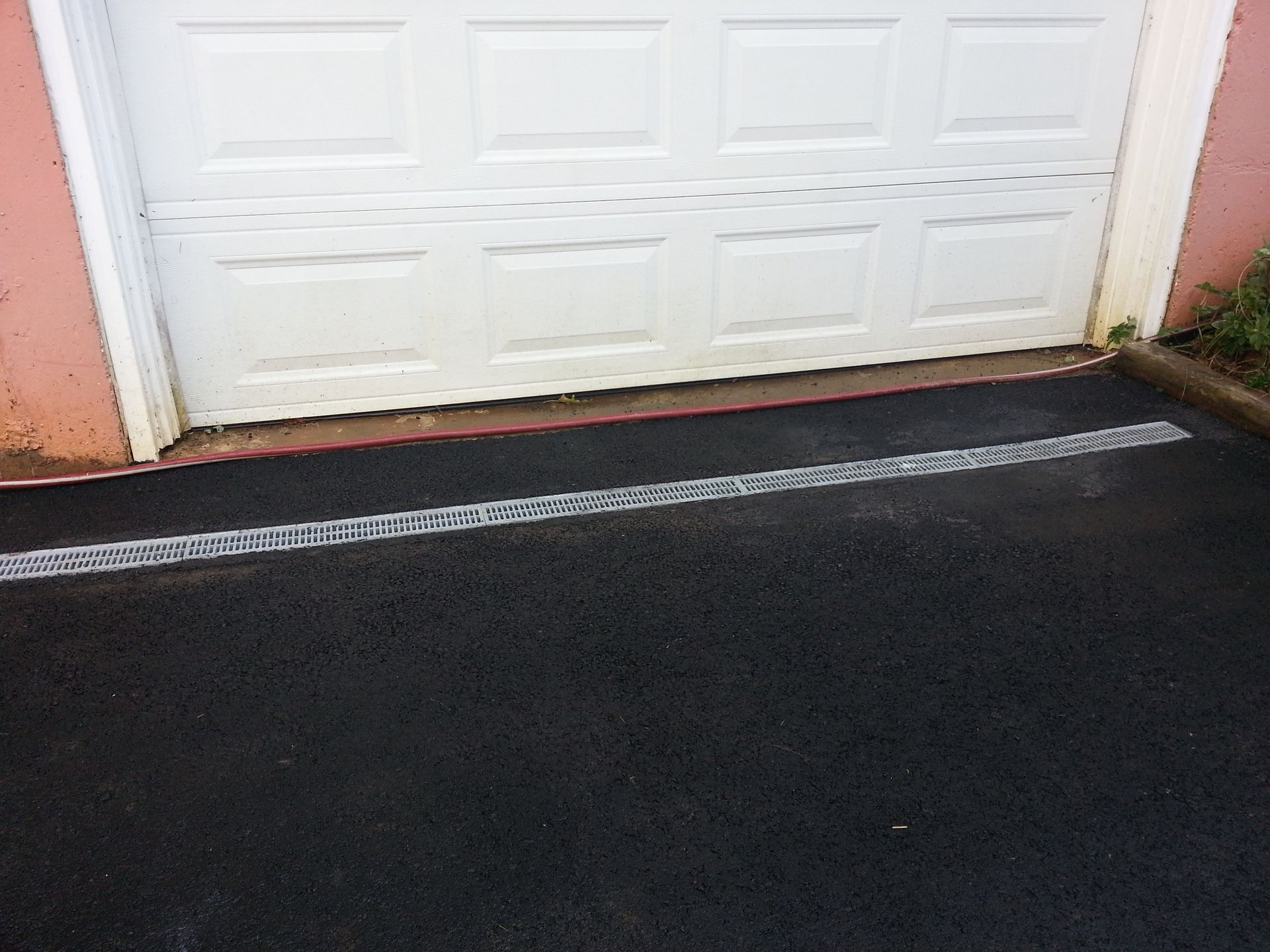 A trench drain with a metal grate installed in an asphalt driveway in front of a white residential garage door.