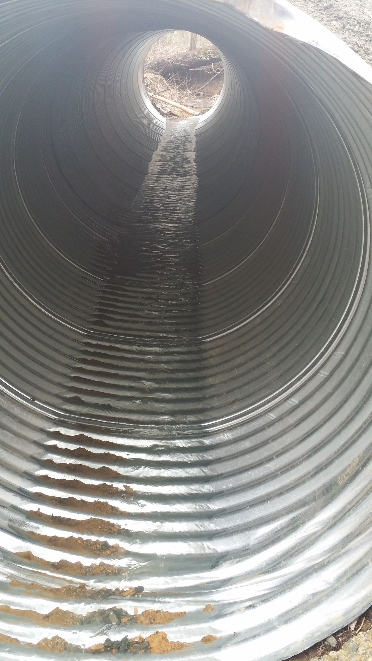 A view through a corrugated metal drainage pipe, showing a narrow stream of water flowing along the bottom toward daylight.