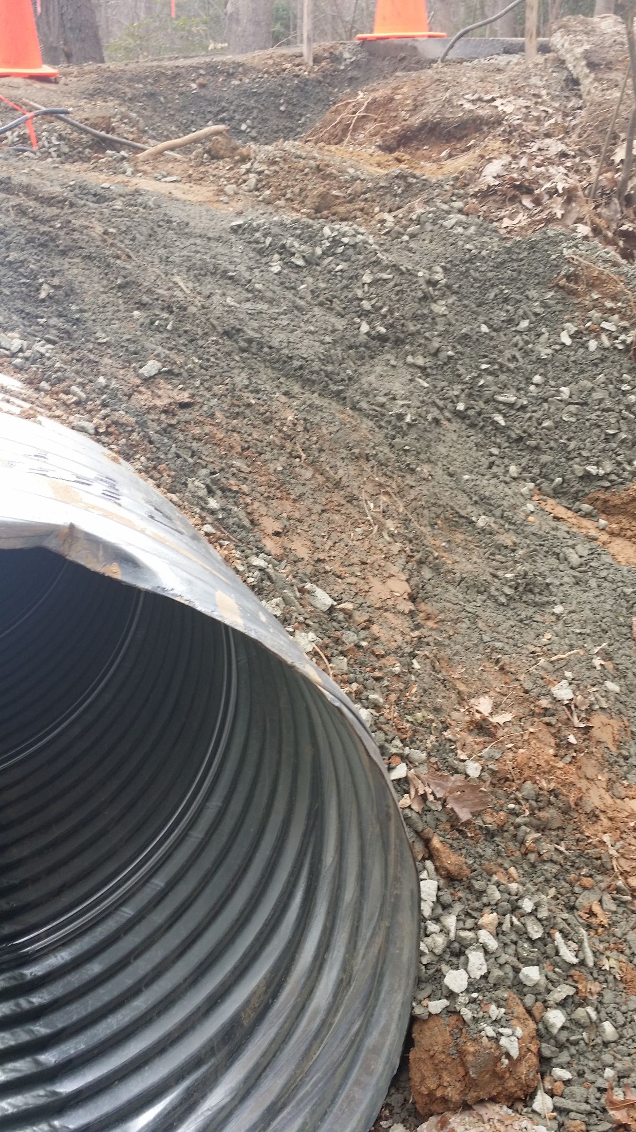 A large, corrugated metal drainage pipe sits at the edge of a gravel-covered construction site with orange cones.