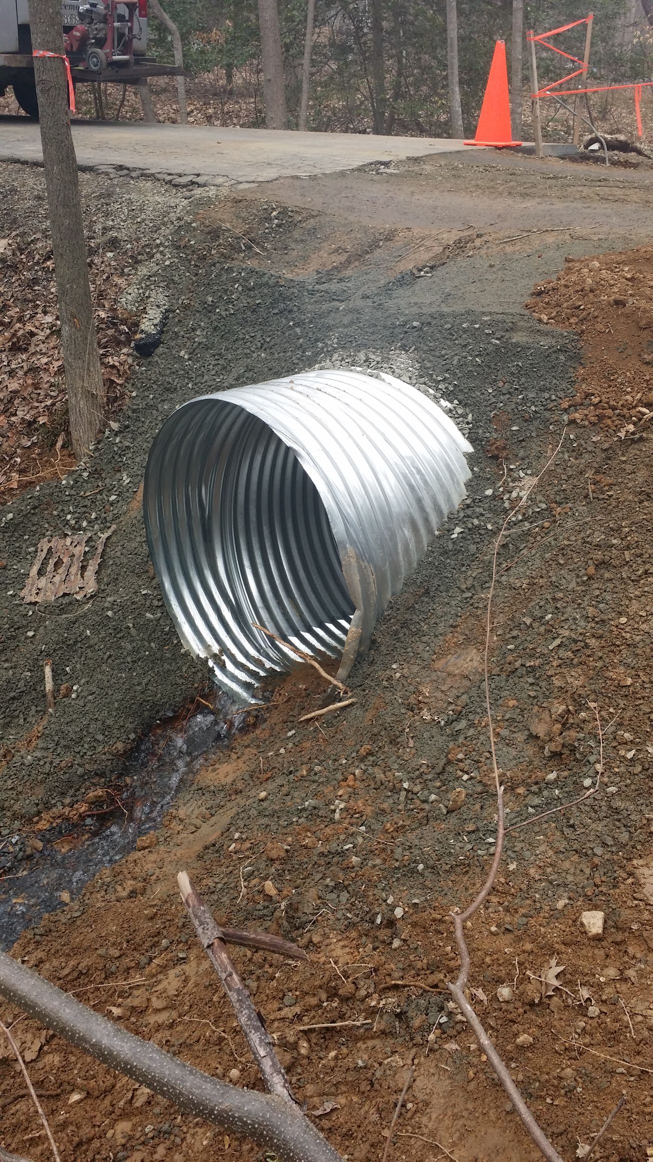 A corrugated metal culvert pipe sits embedded in a gravel embankment with water flowing out of it onto a dirt path.
