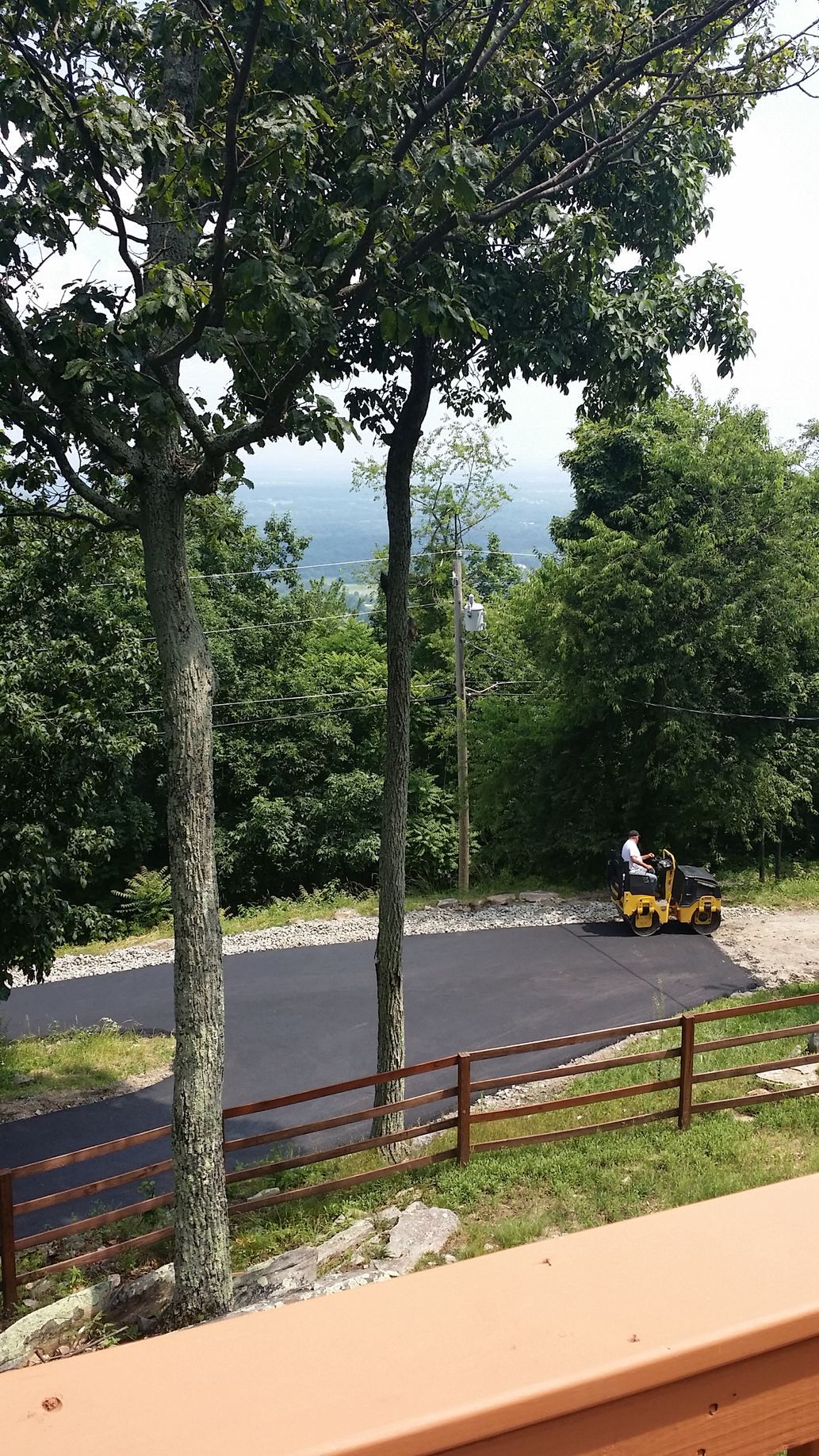 A yellow roller works on a newly paved asphalt driveway overlooking a wooded mountain view behind a wooden fence.