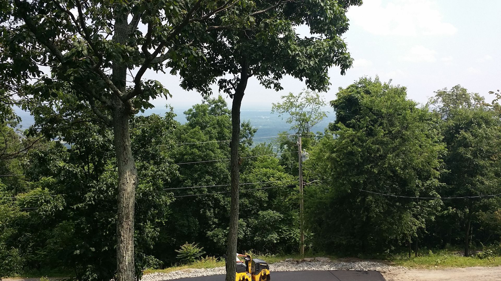 A yellow construction vehicle sits on a paved surface at the edge of a lush, wooded overlook on a sunny day.