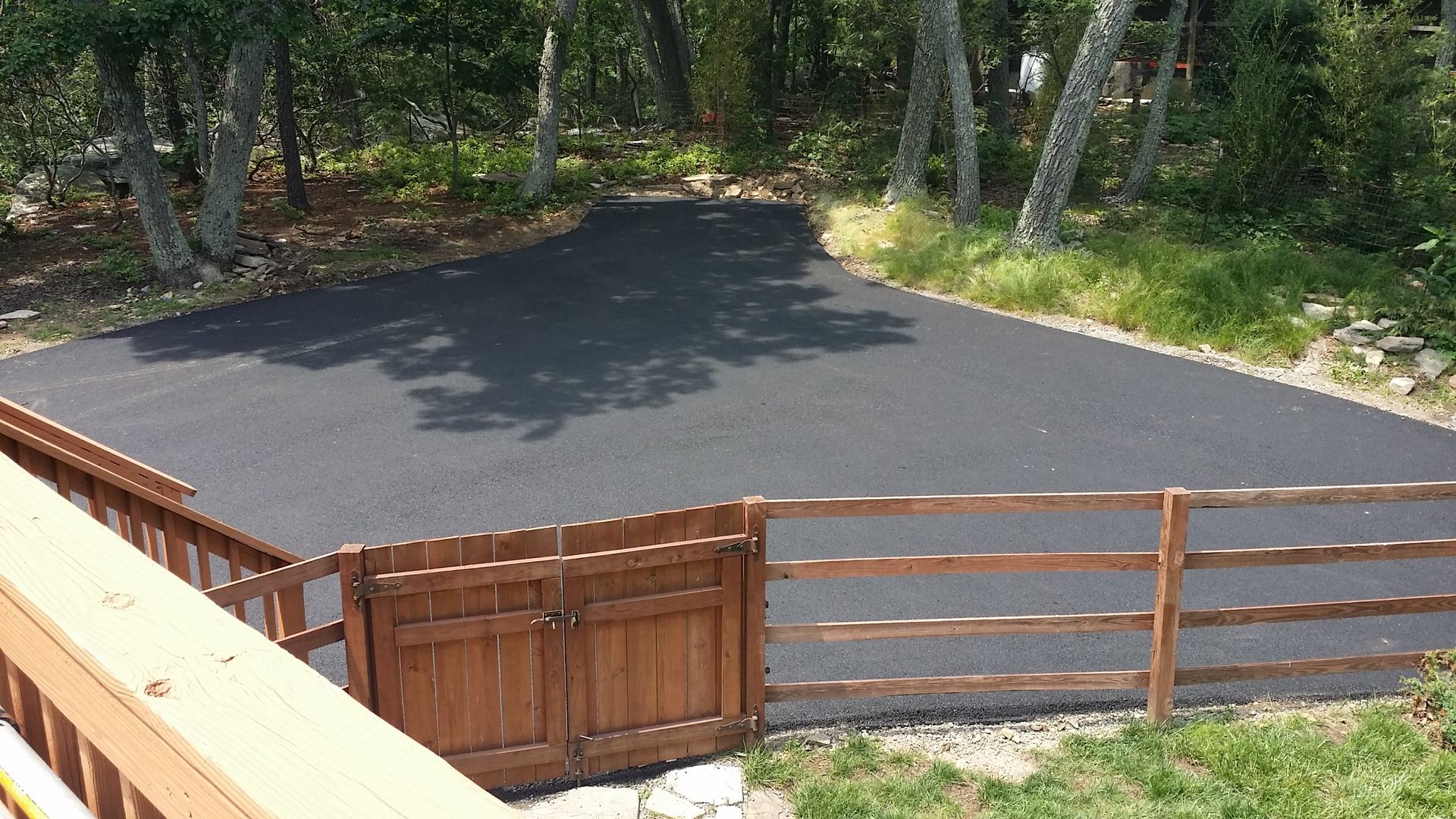 A freshly paved black asphalt driveway enclosed by a low wooden fence and gate, viewed from an elevated deck.