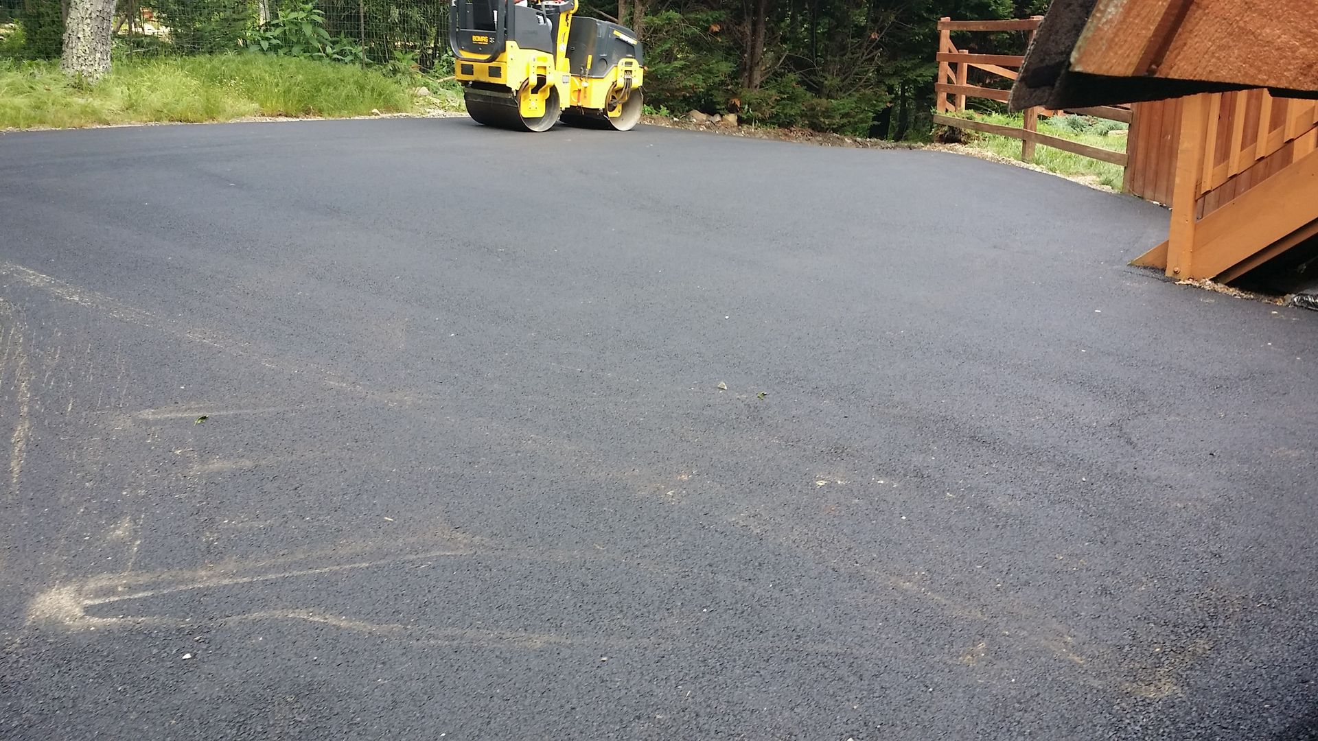 A yellow road roller sits on a newly paved dark asphalt driveway next to wooden stairs and a railing.