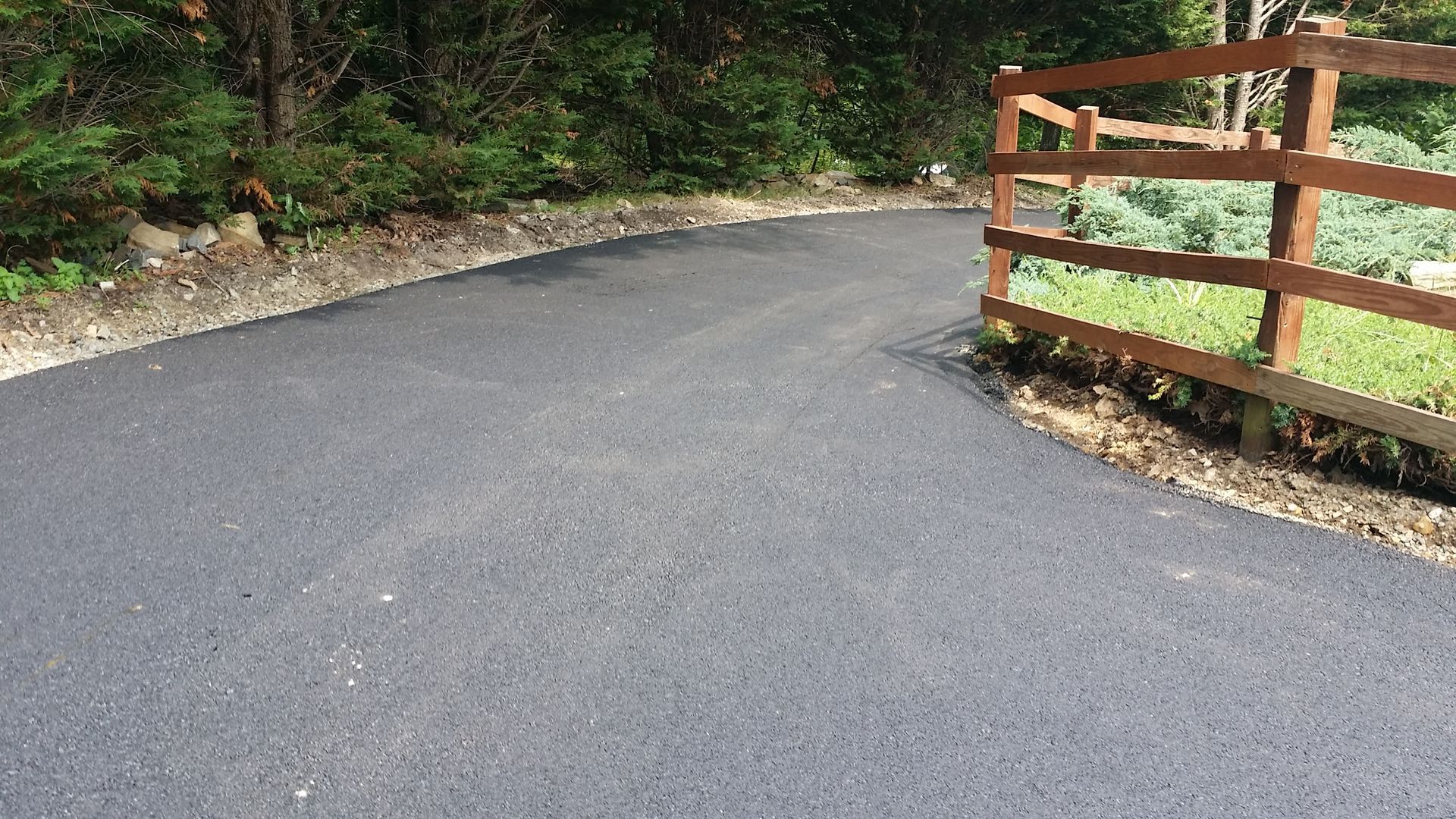A fresh, dark asphalt driveway curves past a wooden post-and-rail fence bordering a landscaped garden.