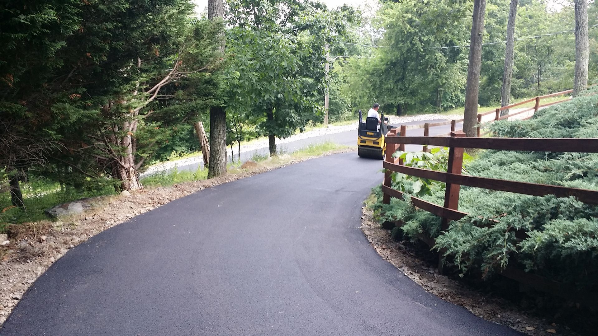 A person drives a yellow paving roller down a freshly laid, winding blacktop driveway bordered by trees and a wooden fence.