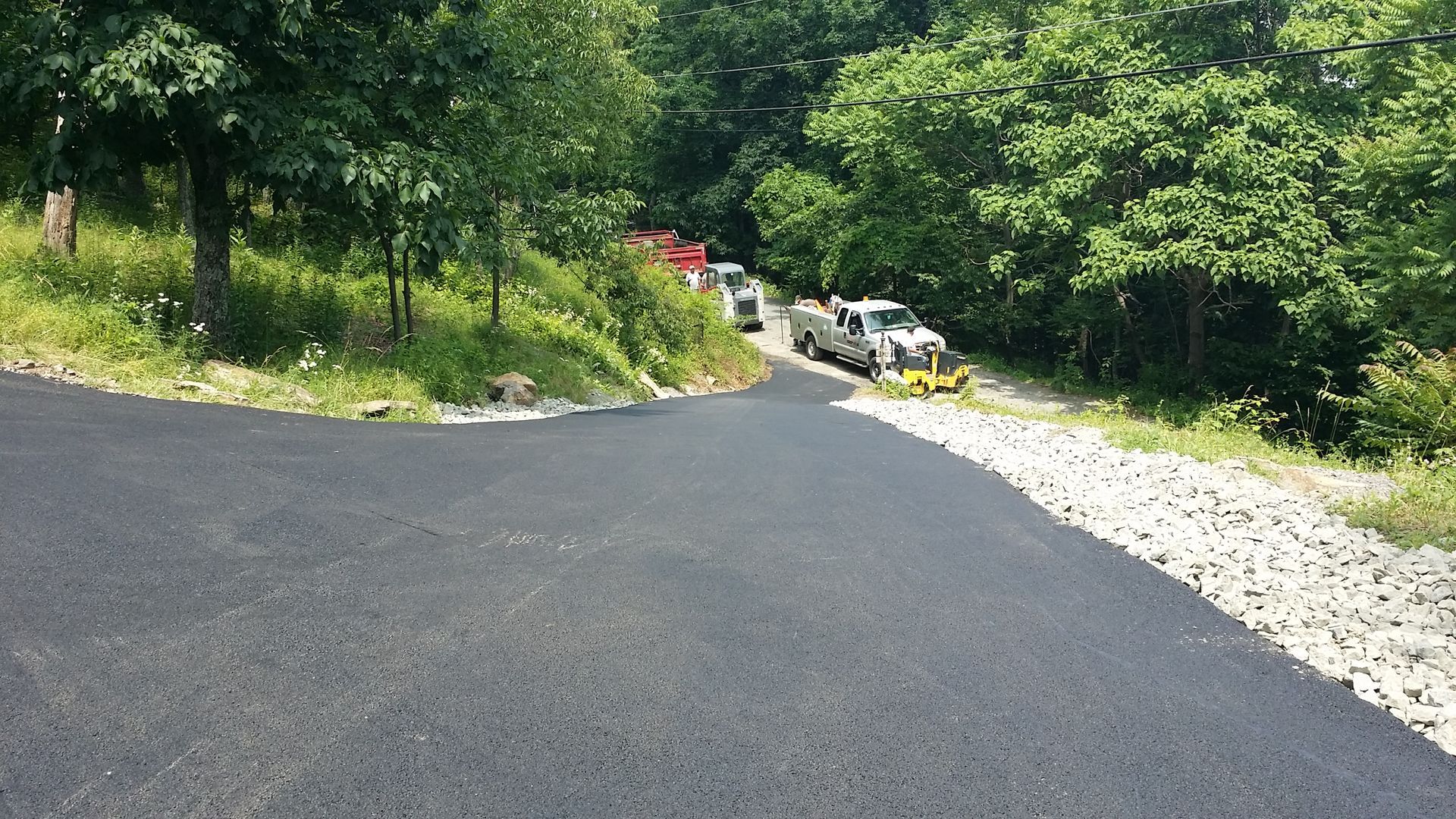 A freshly paved black asphalt driveway curves into a wooded area where a construction truck and equipment are parked.