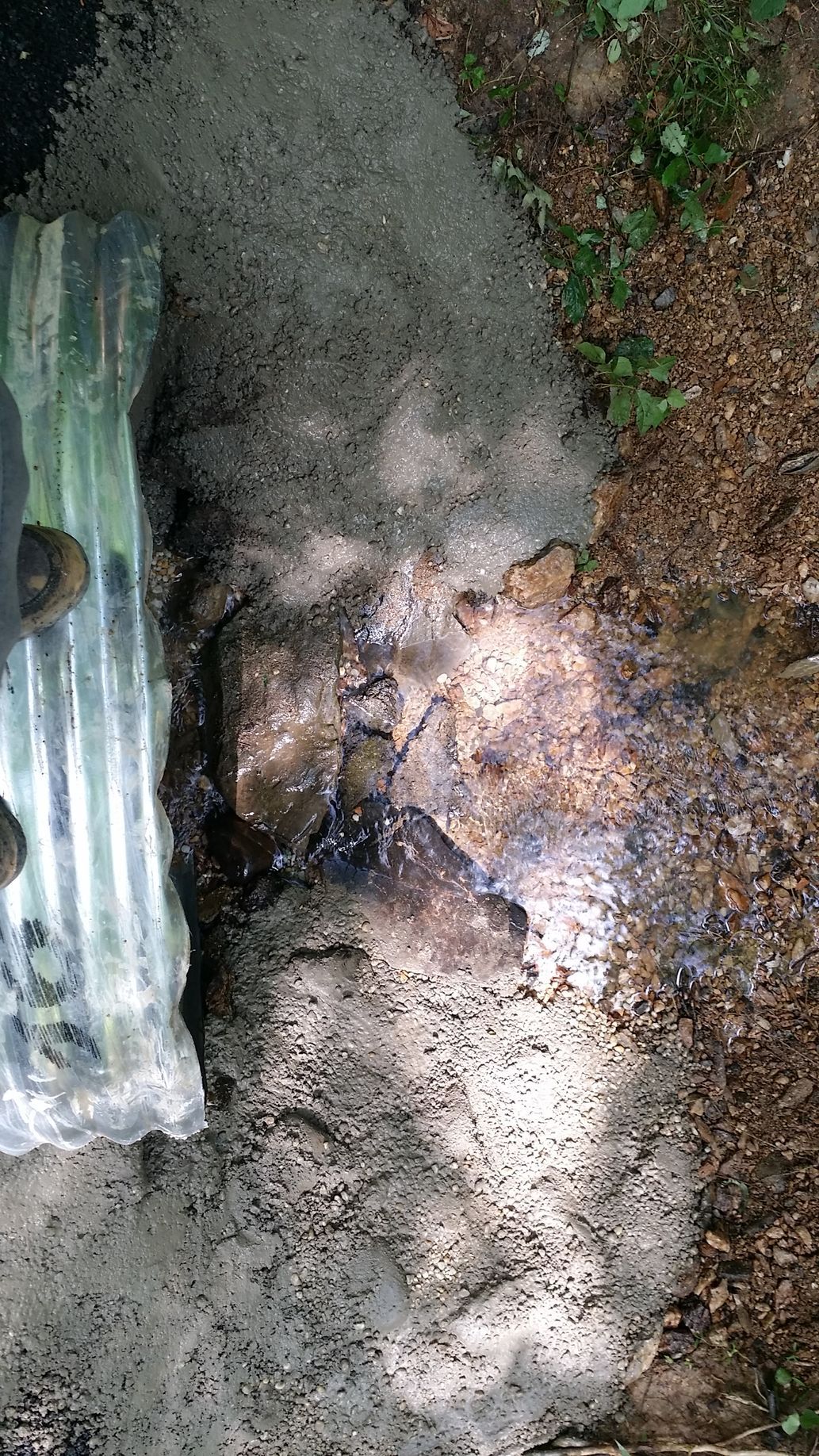 A close-up view of a metal corrugated pipe discharging water onto a patch of dirt and gravel near vegetation.