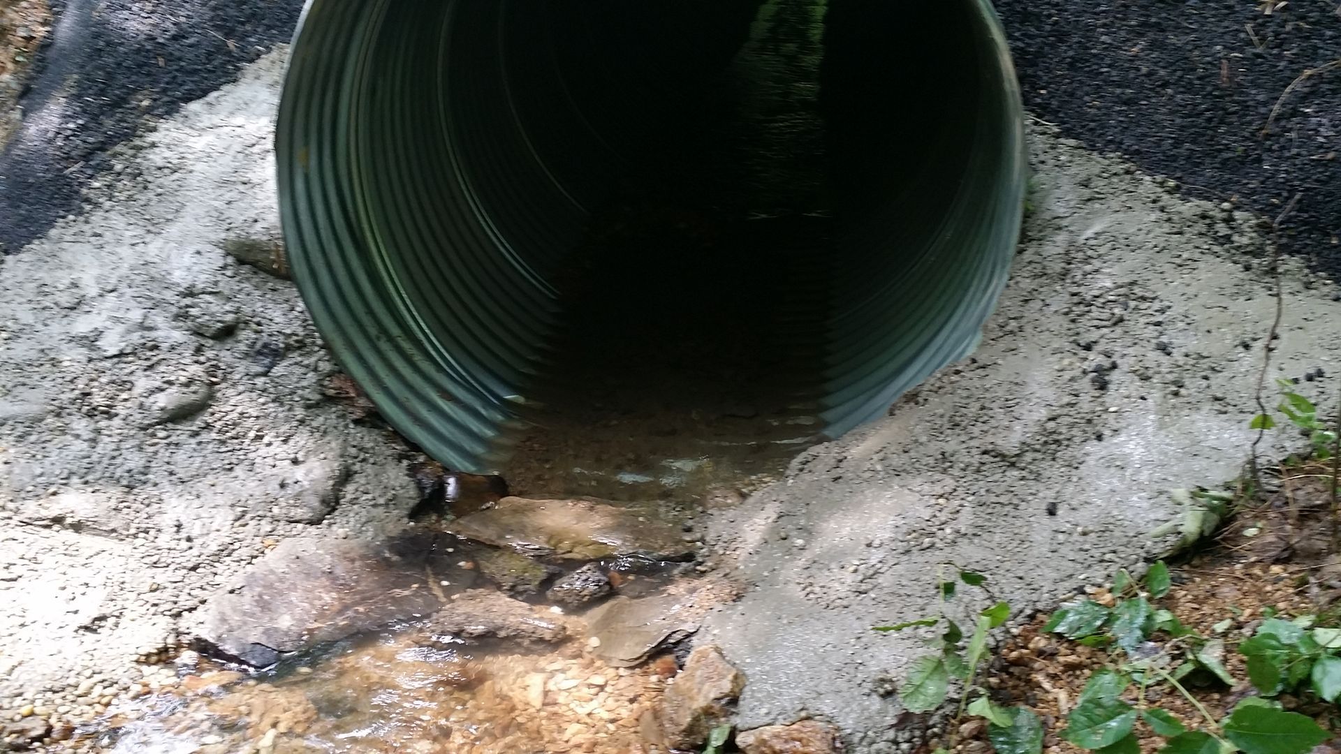 A corrugated metal culvert pipe set into a concrete embankment with a small stream of water flowing out.