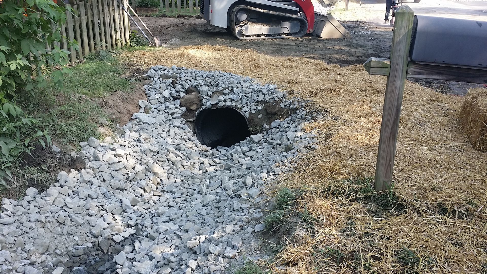 A black culvert pipe sits in a shallow ditch lined with gray crushed stone, with a construction vehicle in the background.