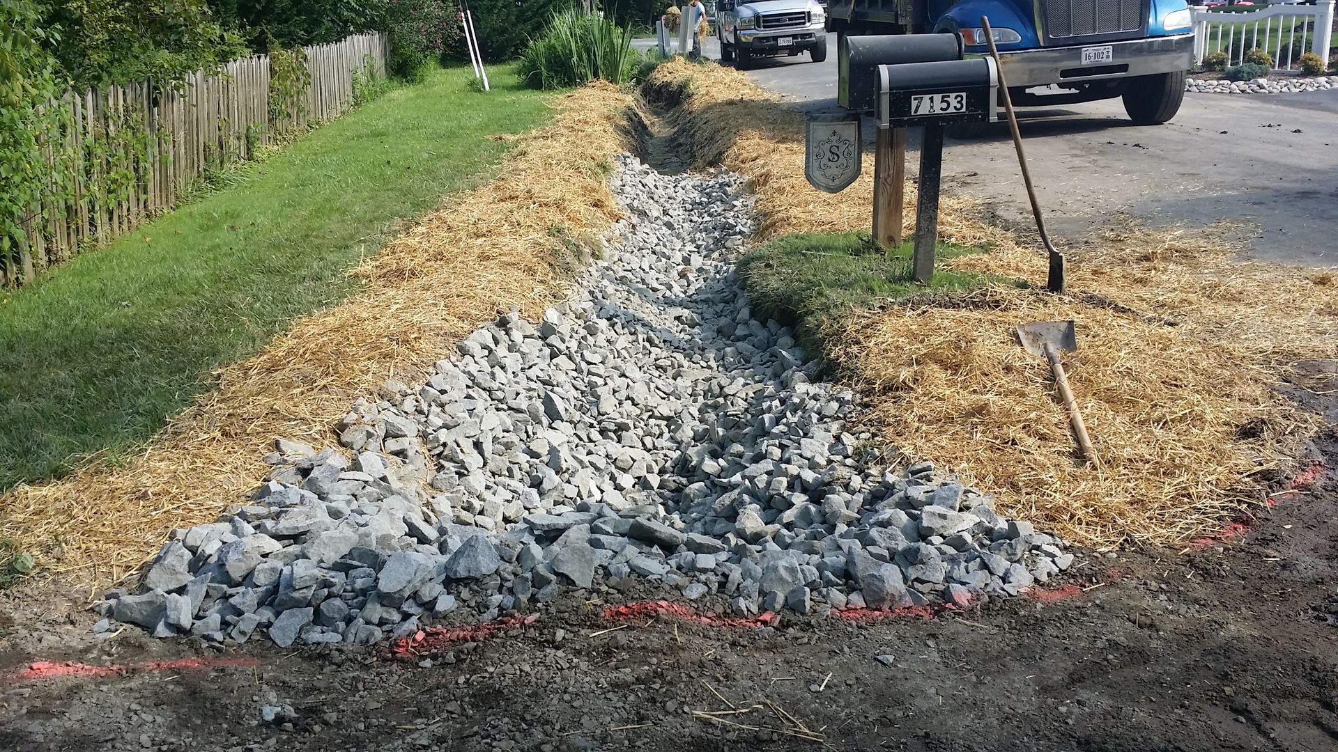 A newly dug drainage ditch lined with grey rocks, surrounded by straw mulch along a residential roadside.