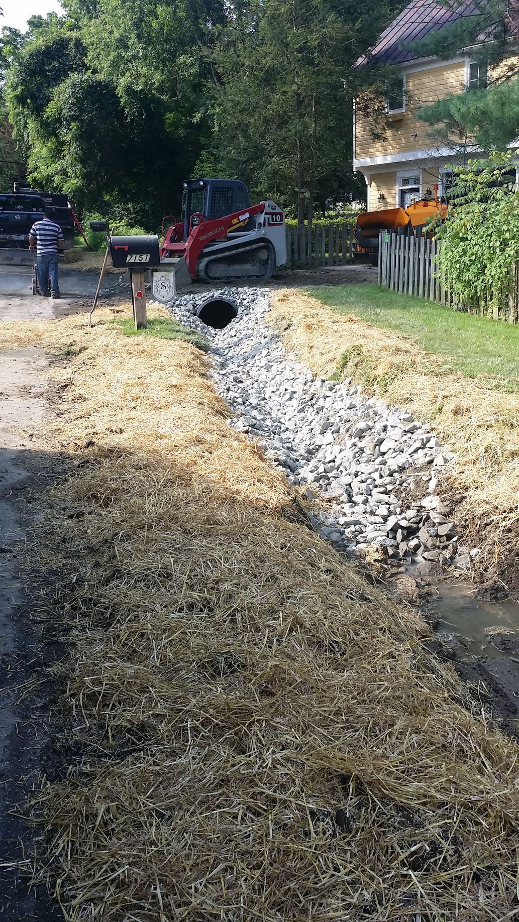 A rock-lined drainage ditch with fresh straw cover runs alongside a road in front of a house, with a skid steer parked.
