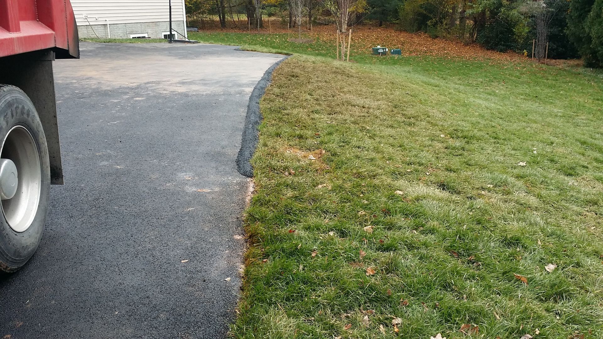 A close-up view of an asphalt driveway edge meeting a slightly sloped green lawn with a black rubber curb guard installed.