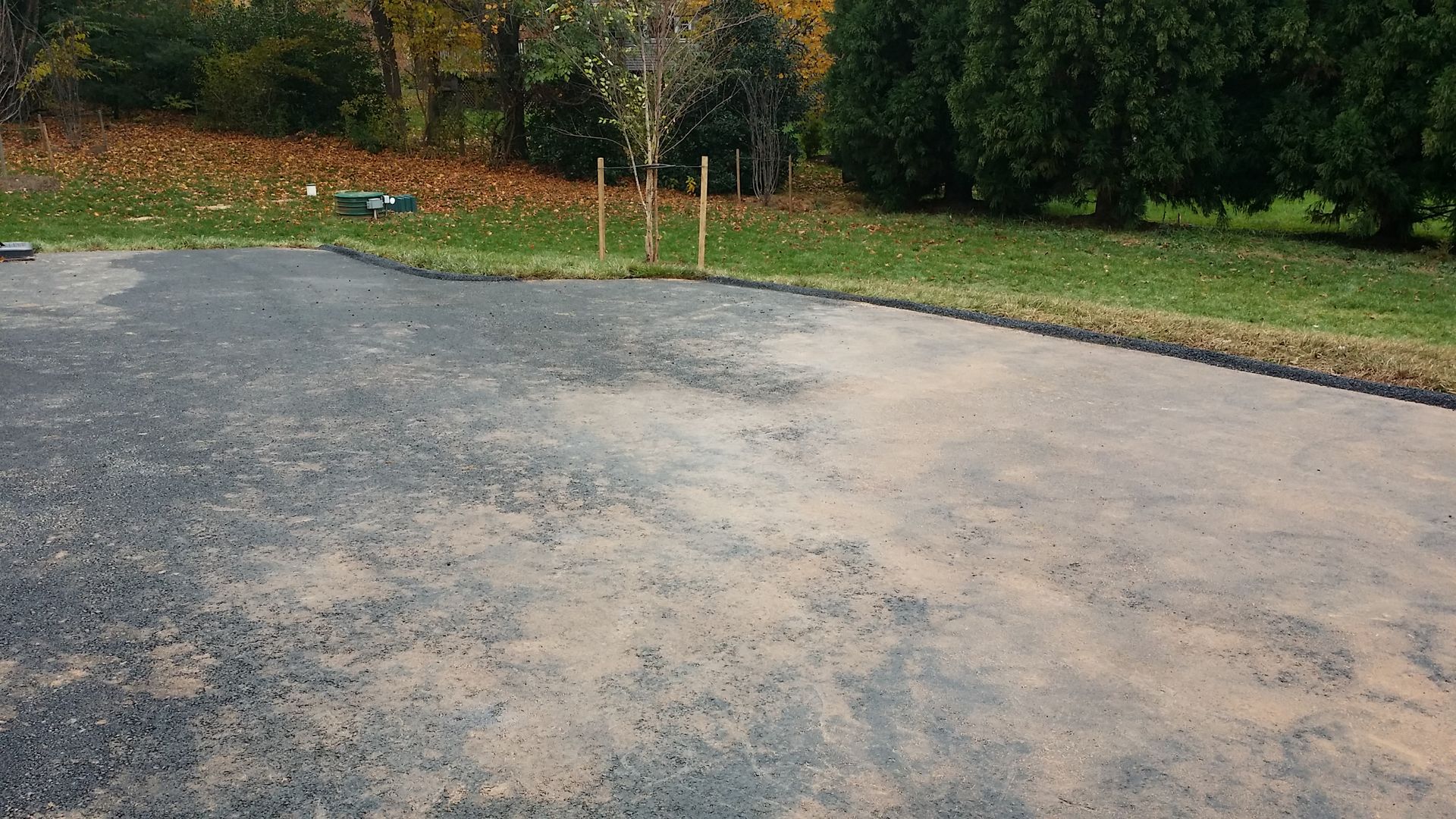 A driveway partially covered in fine gravel or dust, bordering a green lawn and trees in a residential outdoor setting.