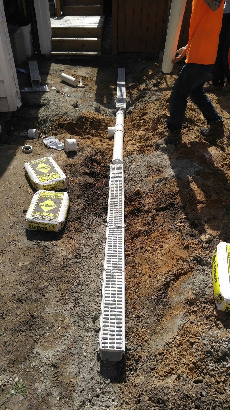 A construction worker installs a trench drain and PVC pipe in a dirt yard next to bags of concrete mix.