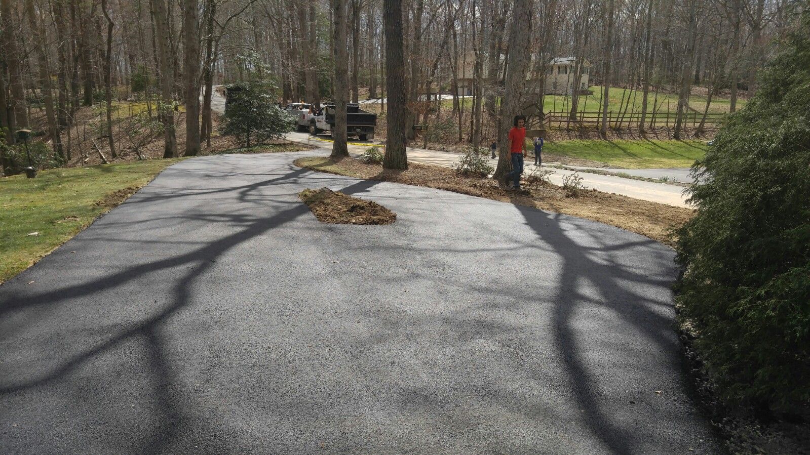 A newly paved asphalt driveway leads toward a wooded area with trees and a parked vehicle in the background.