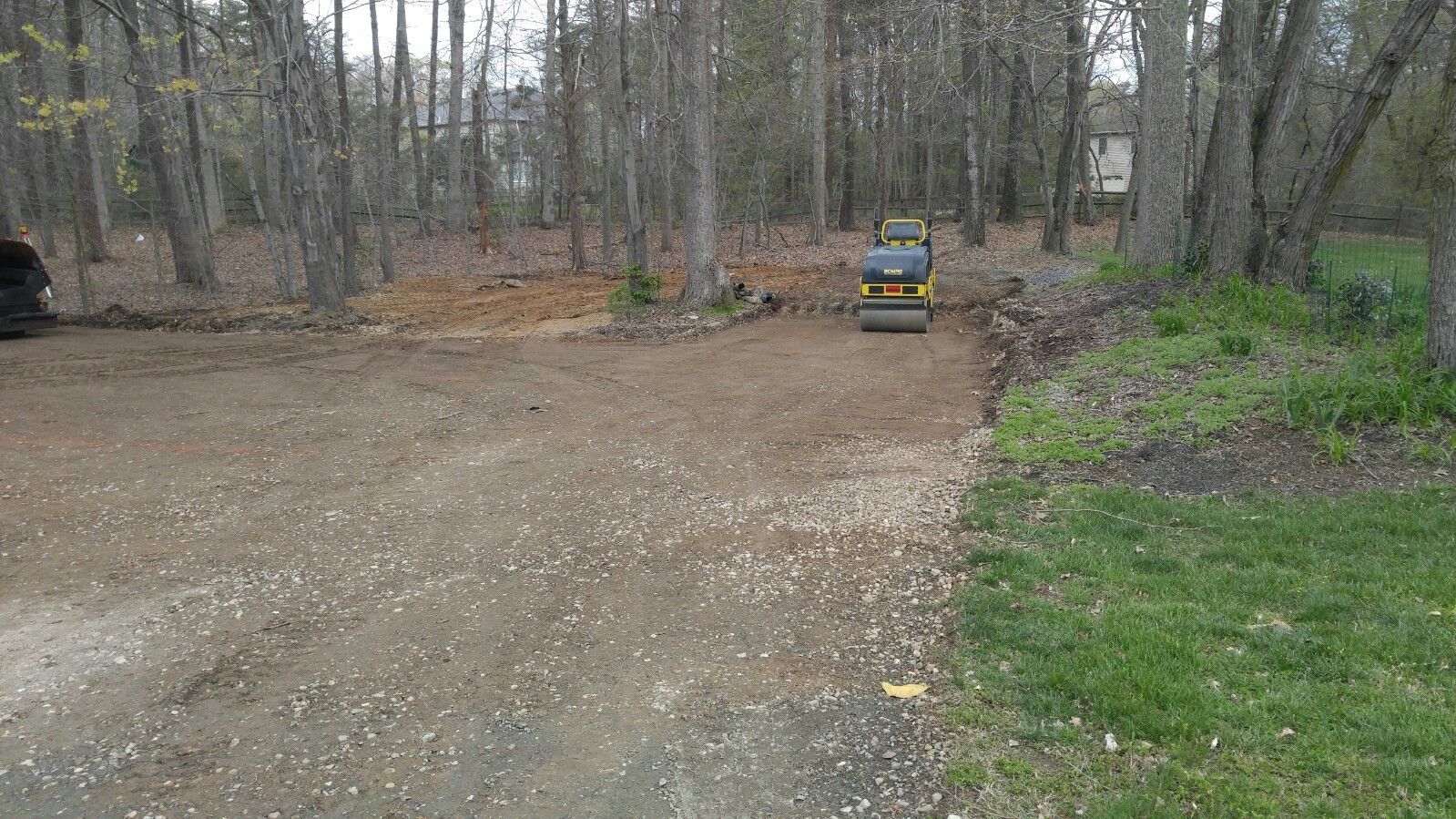 A yellow soil compactor sits on a gravel driveway in a wooded area, grading the ground near a patch of excavated dirt.