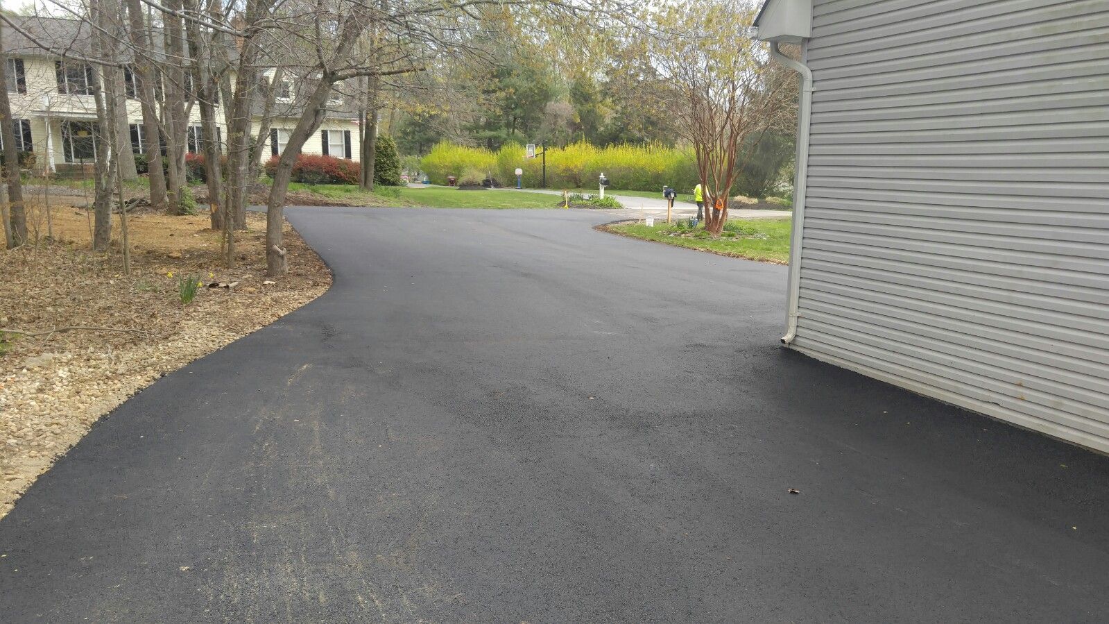 A freshly paved black asphalt driveway leads toward a house corner, with trees and a yellow-flowering bush in the background.