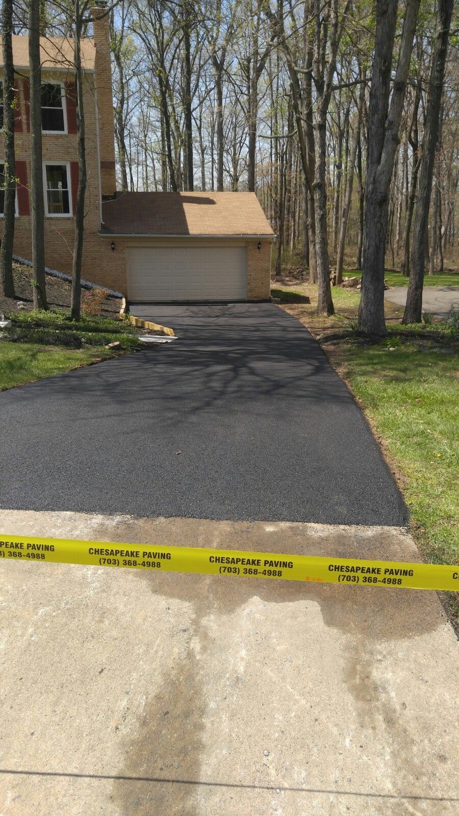 A freshly paved black asphalt driveway leads up to a brick house garage, blocked off by yellow caution tape in the foreground.