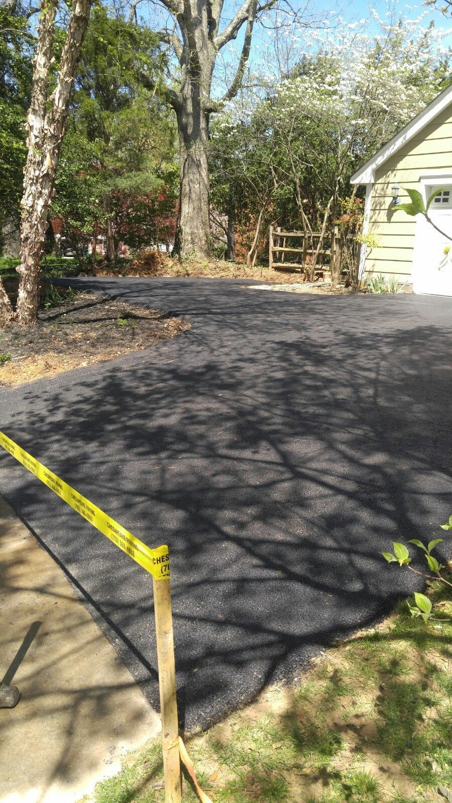 A newly paved black asphalt driveway adjacent to a house, marked off with yellow caution tape on a wooden stake.