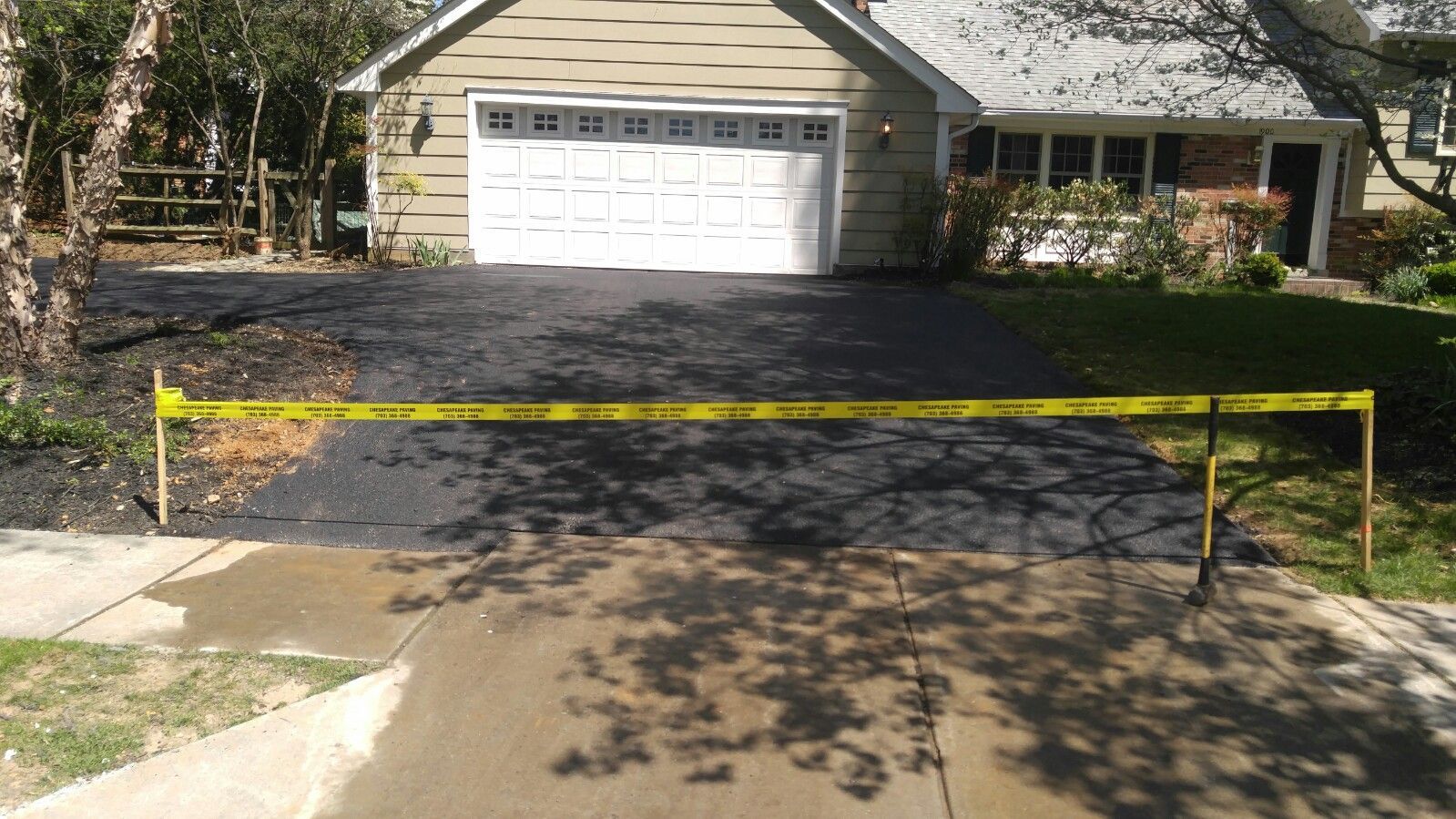 A suburban driveway with fresh black asphalt is blocked off by a horizontal strip of yellow caution tape.