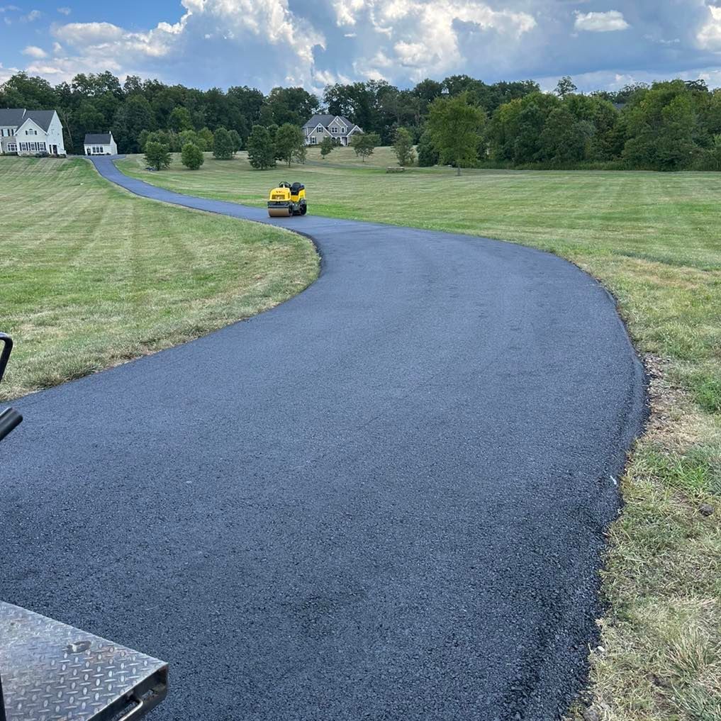 A newly paved, dark asphalt driveway winds through a green grass field toward houses in the distance.